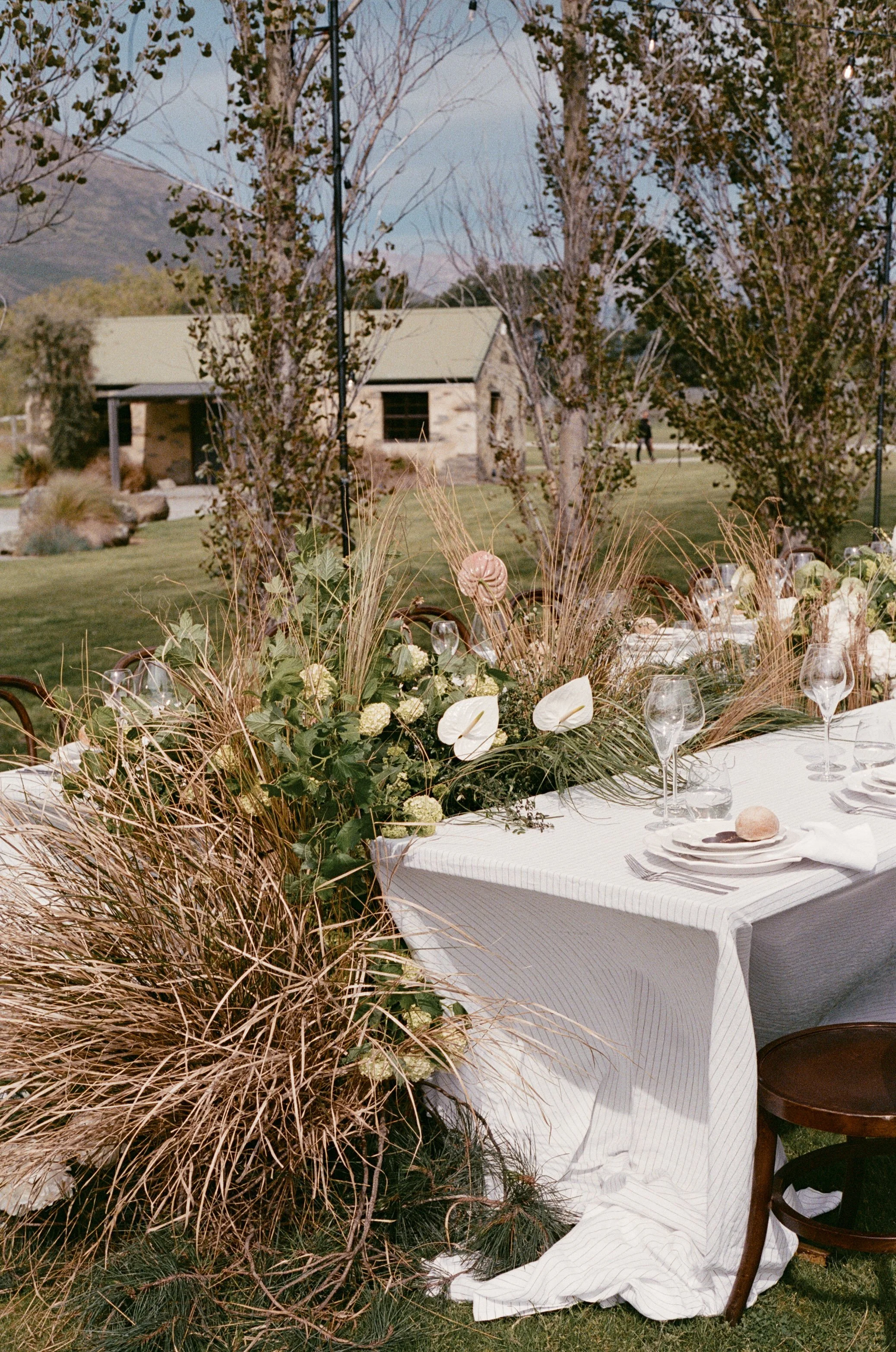 Round outdoor table covered with white tablecloth, set with wine glasses, plates, and silverware, decorated with a large floral arrangement including greenery, white anthuriums, and tall grasses, with trees, a building, and a person in the background.