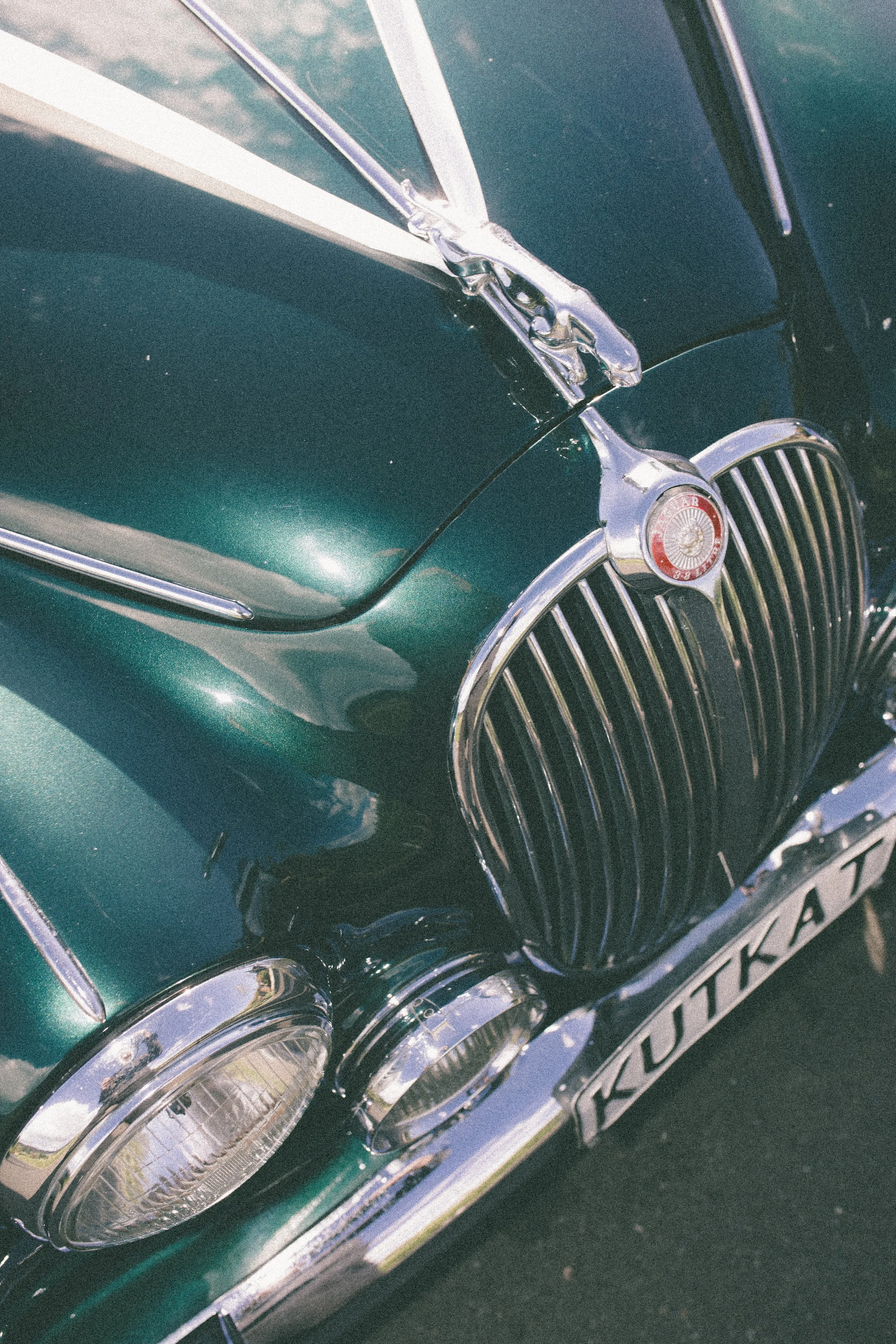 Close-up of the front part of a vintage green Jaguar XK car, showing the grille, headlight, and hood ornament.