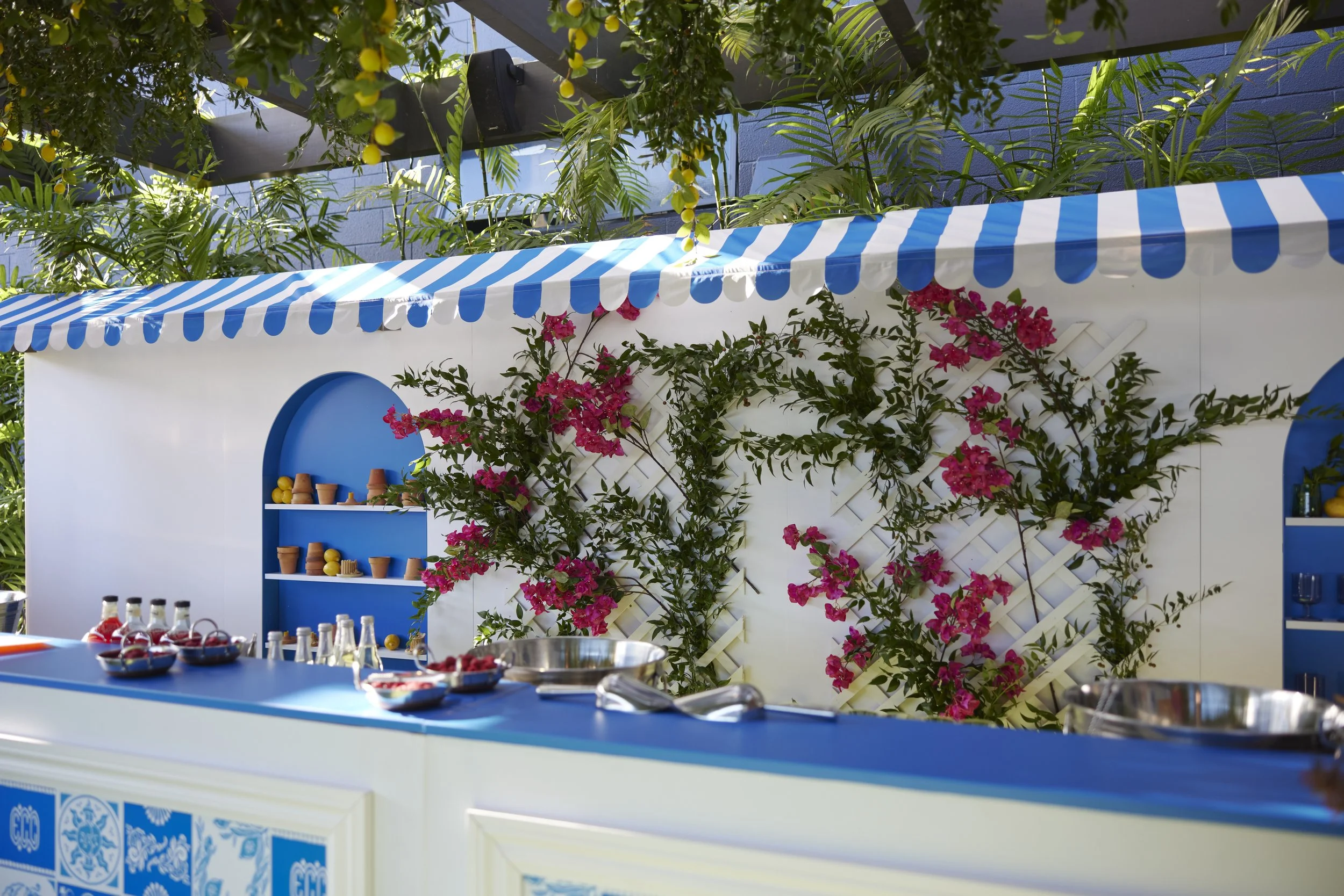 An outdoor bar setup with a blue and white striped canopy, decorated with pink flowers and green foliage. A blue shelf holds yellow lemons and terracotta cups. In the foreground, there is a counter with bowls of berries and bottles.