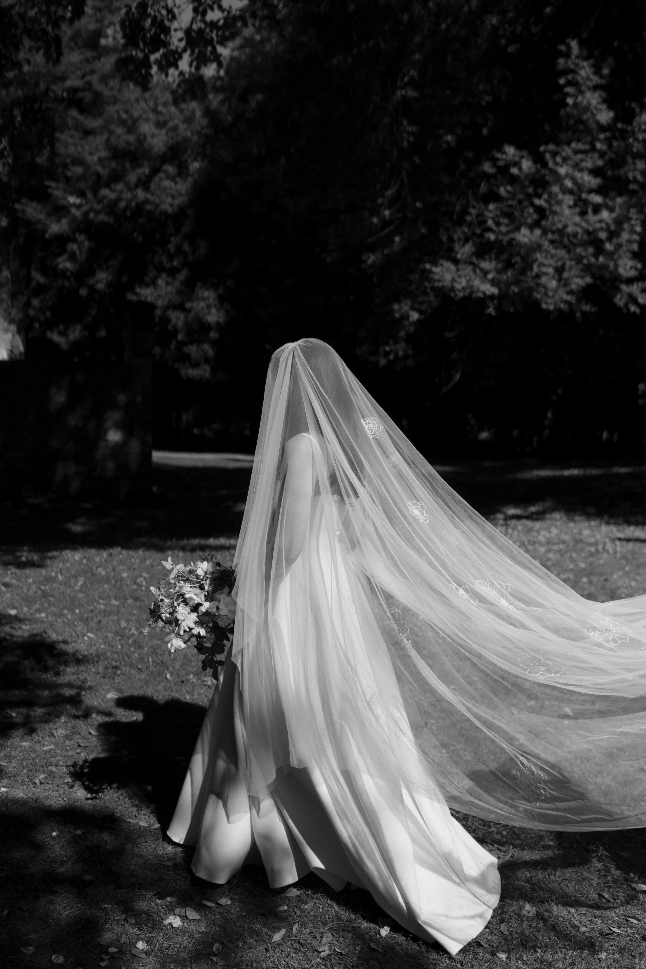 A bride outside in a park, wearing a wedding dress with a long veil covering her face, holding a bouquet of flowers, trees in the background.