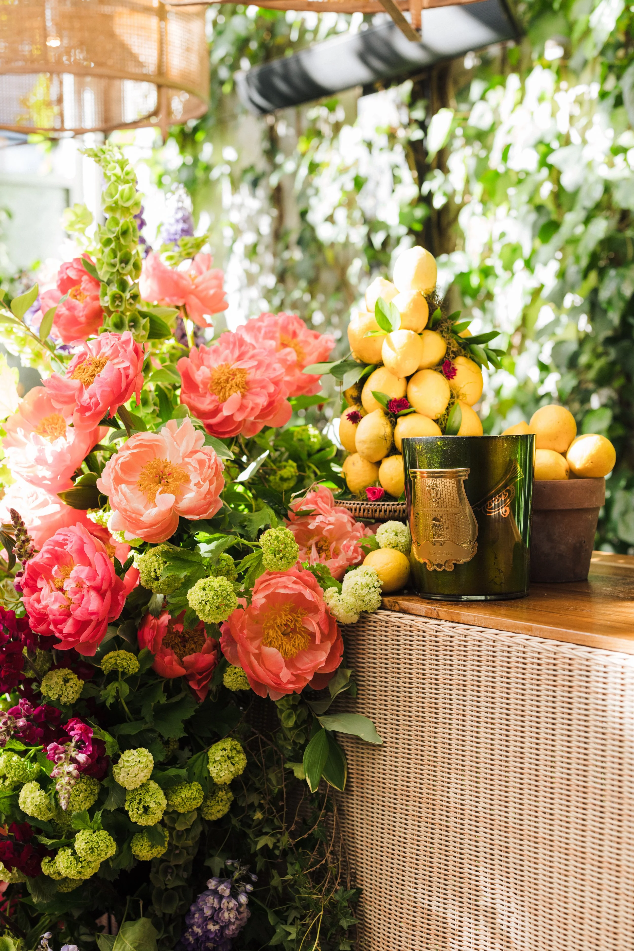 A display of pink flowers with green foliage next to a wicker table holding a stem of yellow lemons, a glass with a dark label, and a potted plant, set in a lush garden.