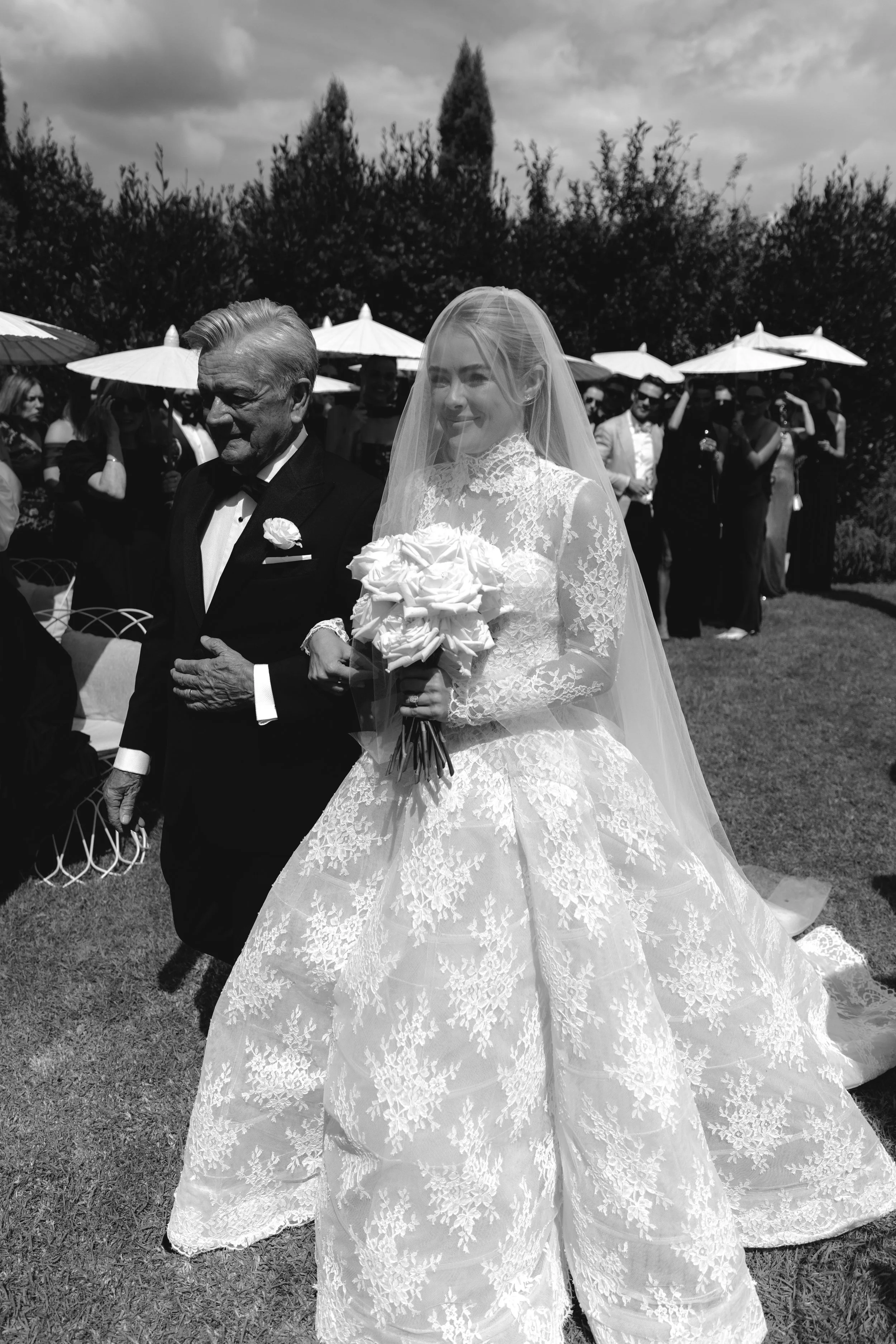 A bride in a lace wedding gown holding a bouquet walking outdoors with an elderly man in a suit, surrounded by friends and family under umbrellas.