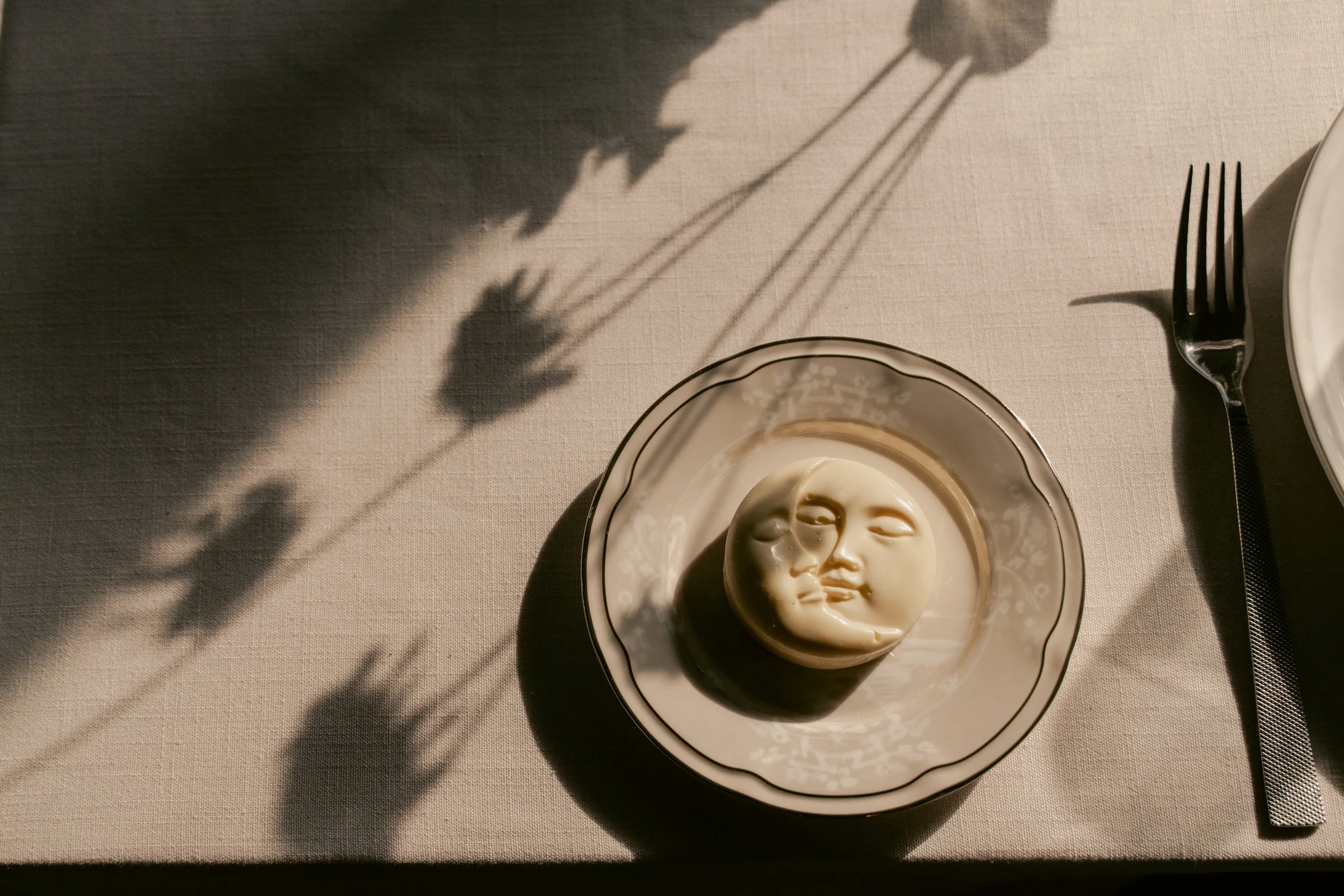 A dish of creamy sauce with a face design, placed on a table with a fork and a plate nearby, with shadows cast on the table.