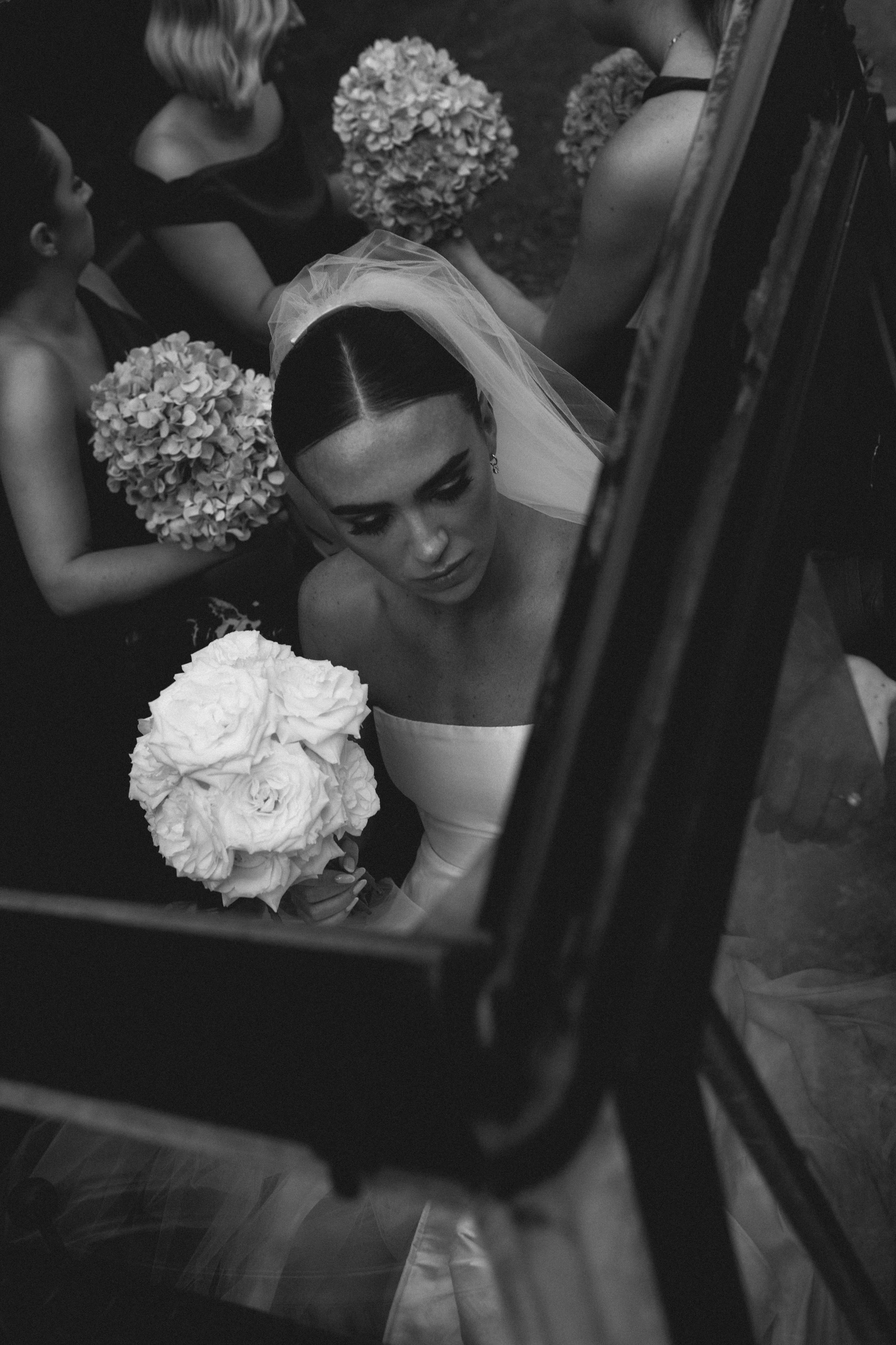 A bride in a white wedding dress with a veil, holding a bouquet of white flowers, is seen through what appears to be a mirror or reflective surface, surrounded by her bridesmaids holding similar bouquets.