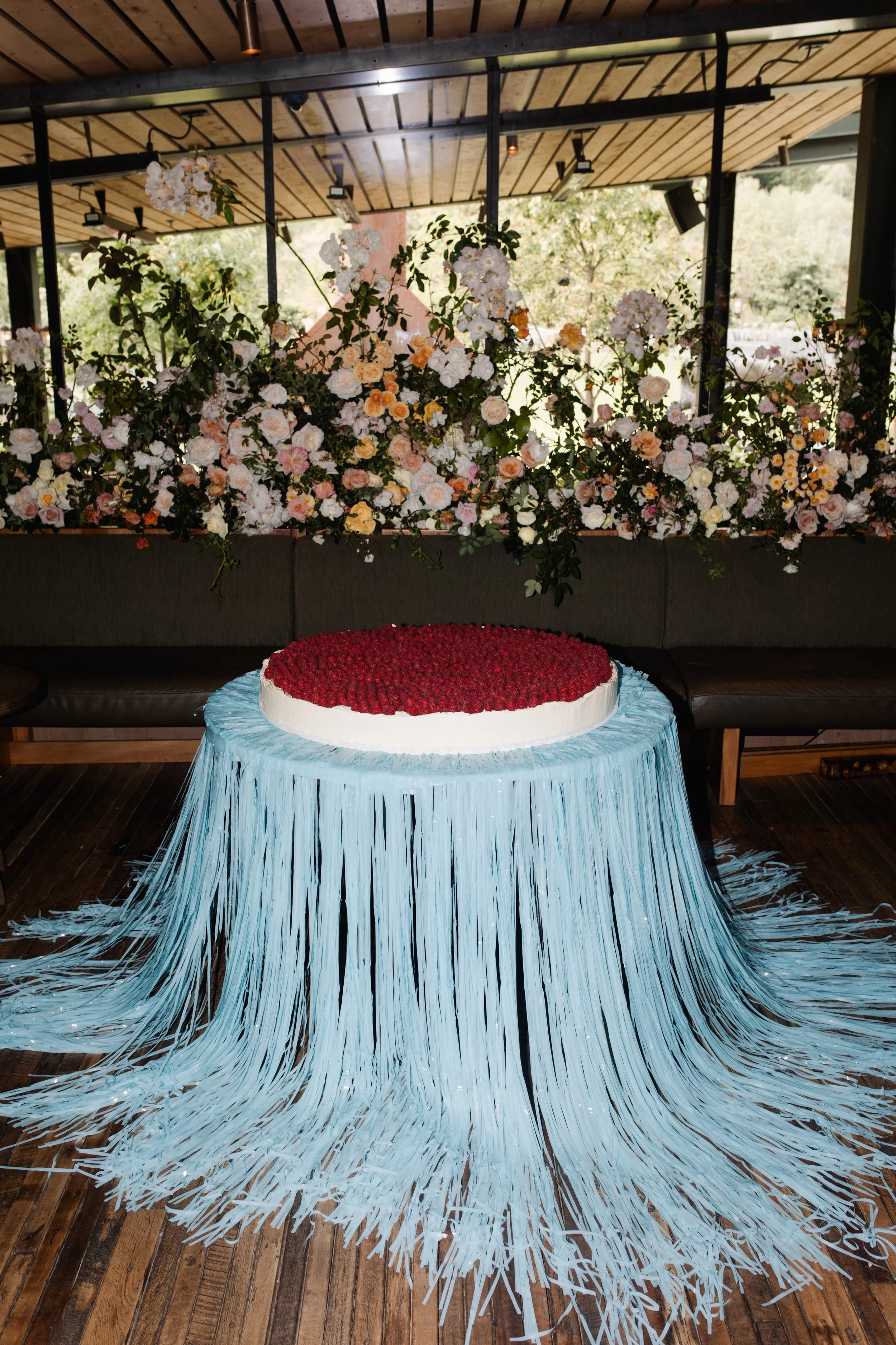 A round cake with a white frosting border and red topping, placed on a stand with blue streamers hanging down.
