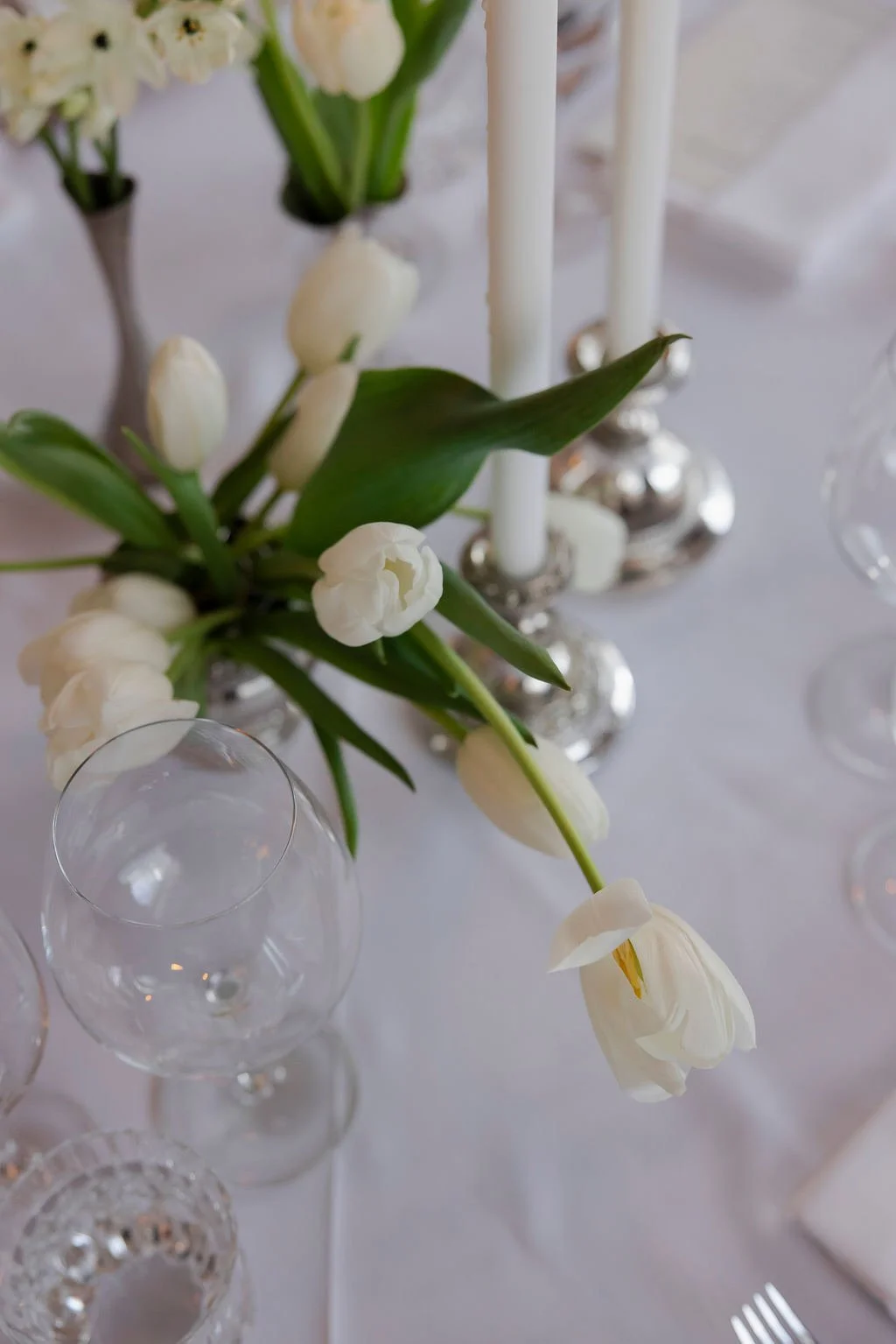 White tulips and flowers in a silver vase on a white table, with empty glassware nearby.