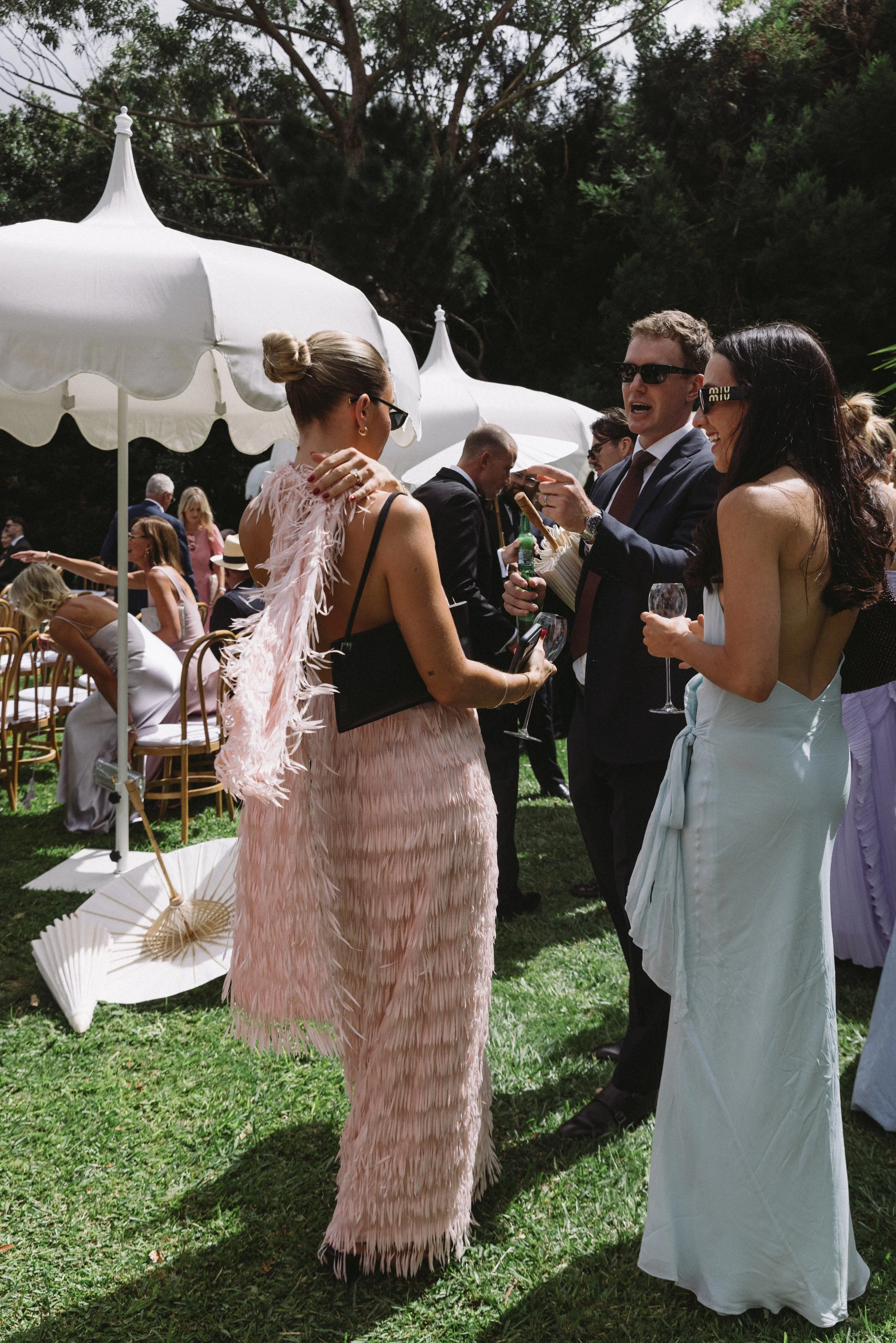 People attending a social gathering outdoors under umbrellas, dressed in formal and semi-formal attire, engaging in conversations and enjoying drinks on a sunny day.