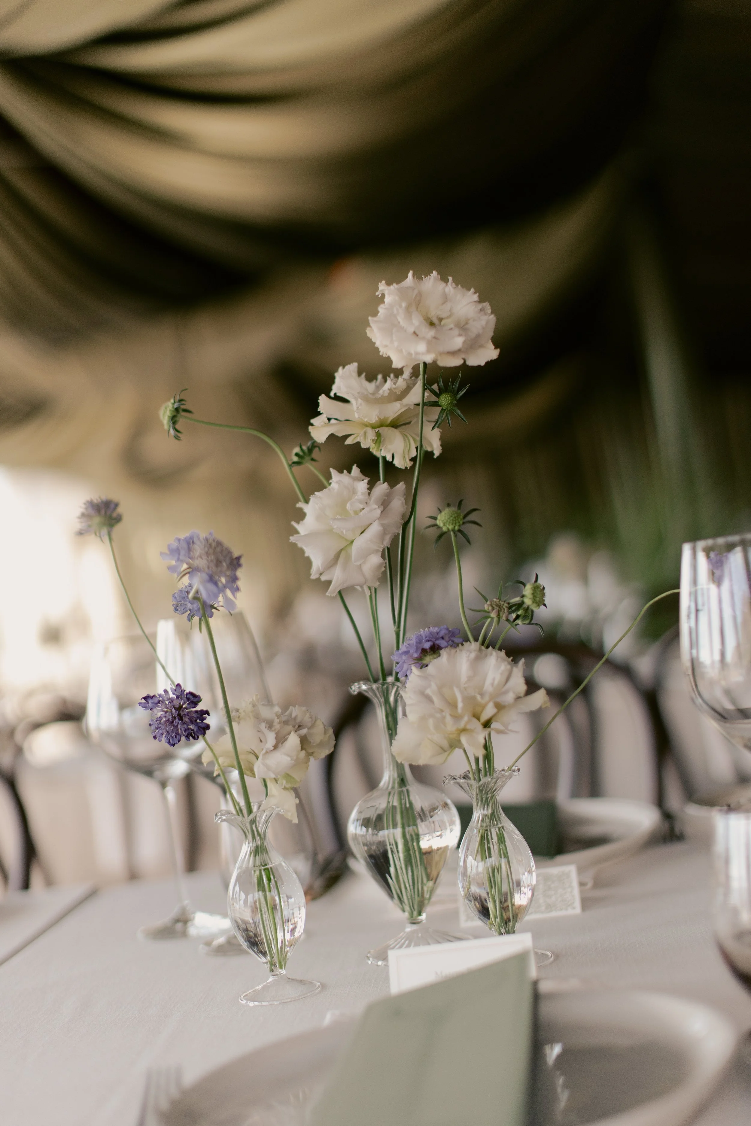 Elegant table centerpiece with white and purple flowers in clear glass vases, set on a white tablecloth with wine glasses and menus. The background is blurred with a dark ceiling or drapery.
