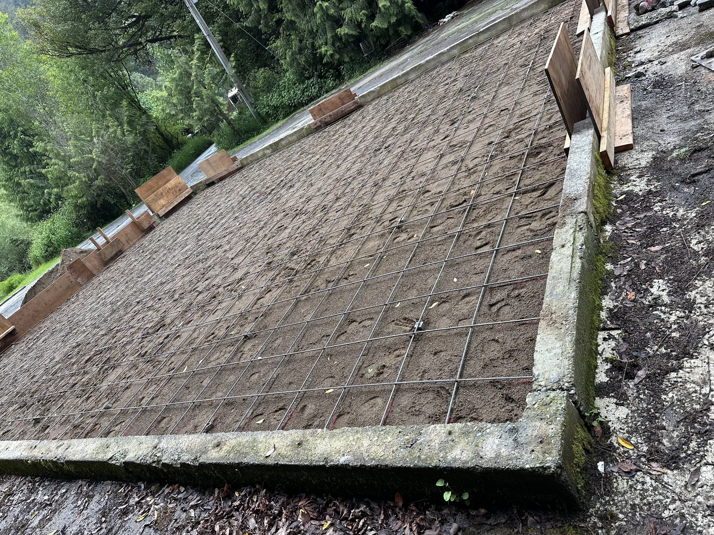 A construction site showing a concrete wall with an exposed wire mesh reinforcement, surrounded by a dirt and gravel ground, with trees and greenery in the background.