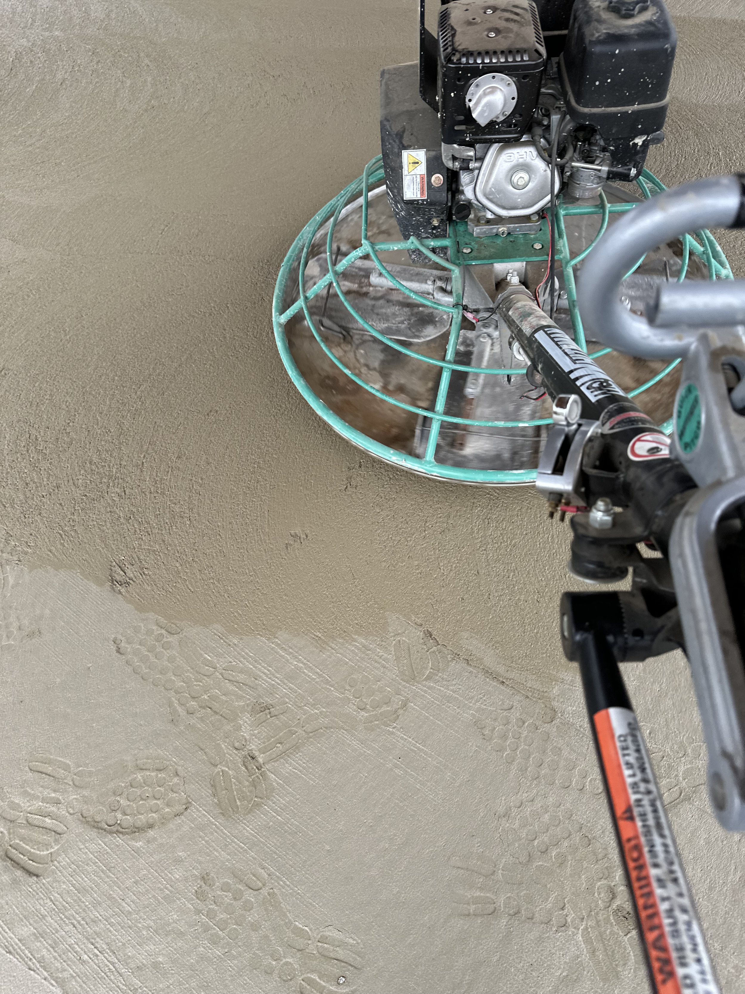 A close-up of a power trowel smoothing freshly poured concrete on a floor, with visible footprints in the concrete.