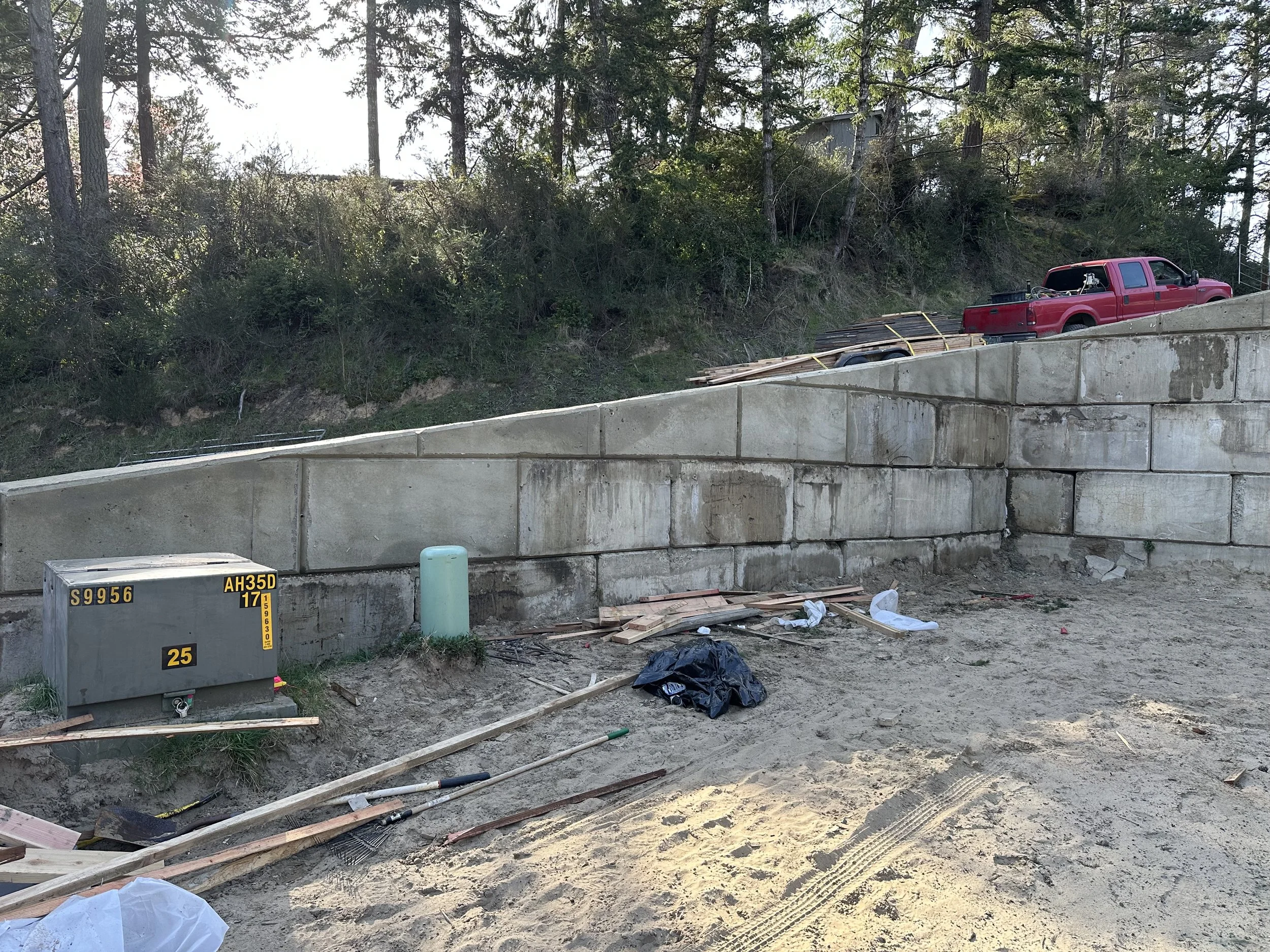 Construction site with a partially built concrete wall, construction tools, and a red pickup truck parked on an embankment among trees and bushes.