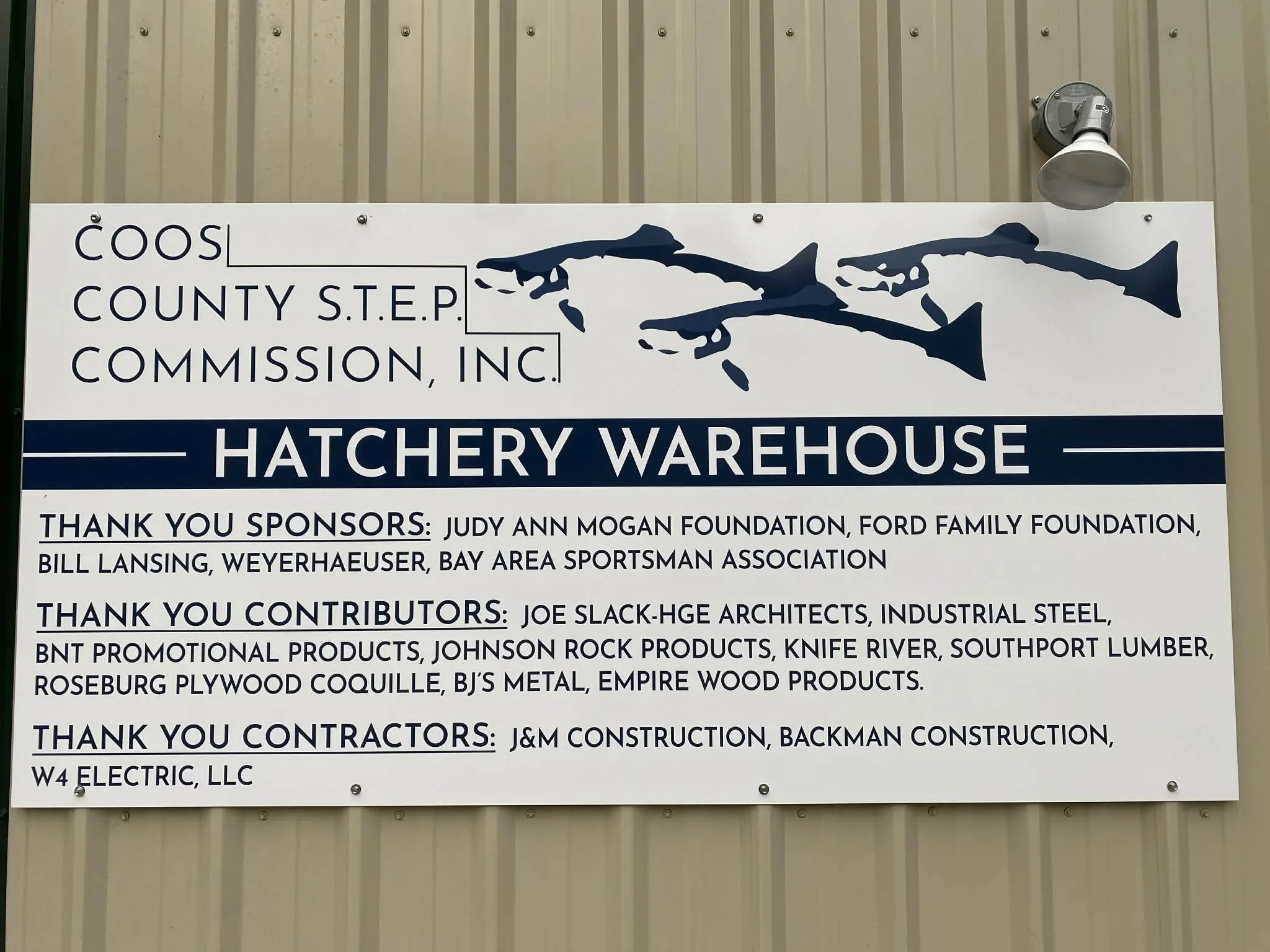 A sign for the Coos County S.T.E.P. Hatchery Warehouse. The sign features the outline of a fish and lists sponsors and contributors for the project.