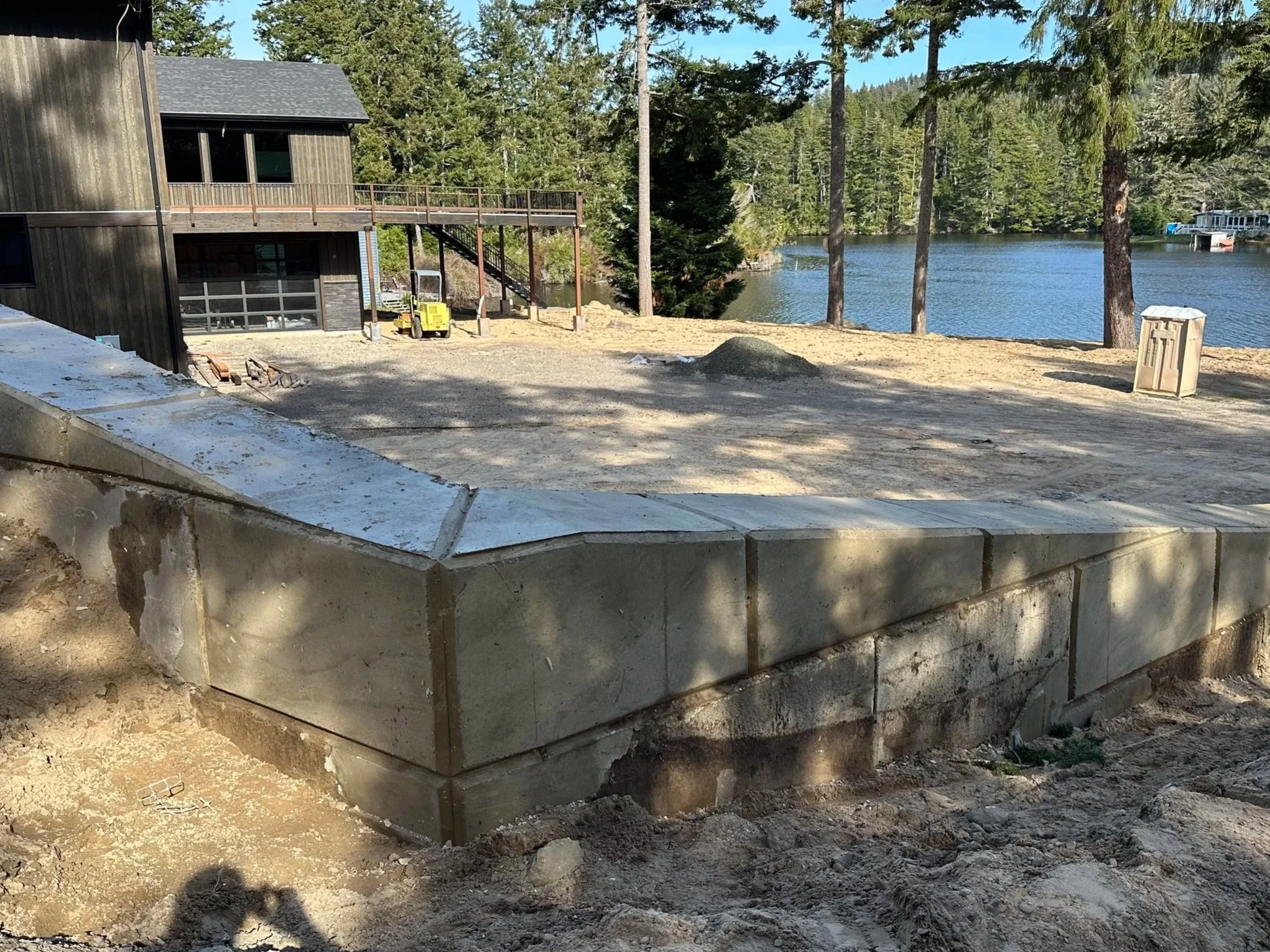 Construction site near a lake with a black wooden building, a gravel area, and a concrete retaining wall, surrounded by tall pine trees and blue sky.