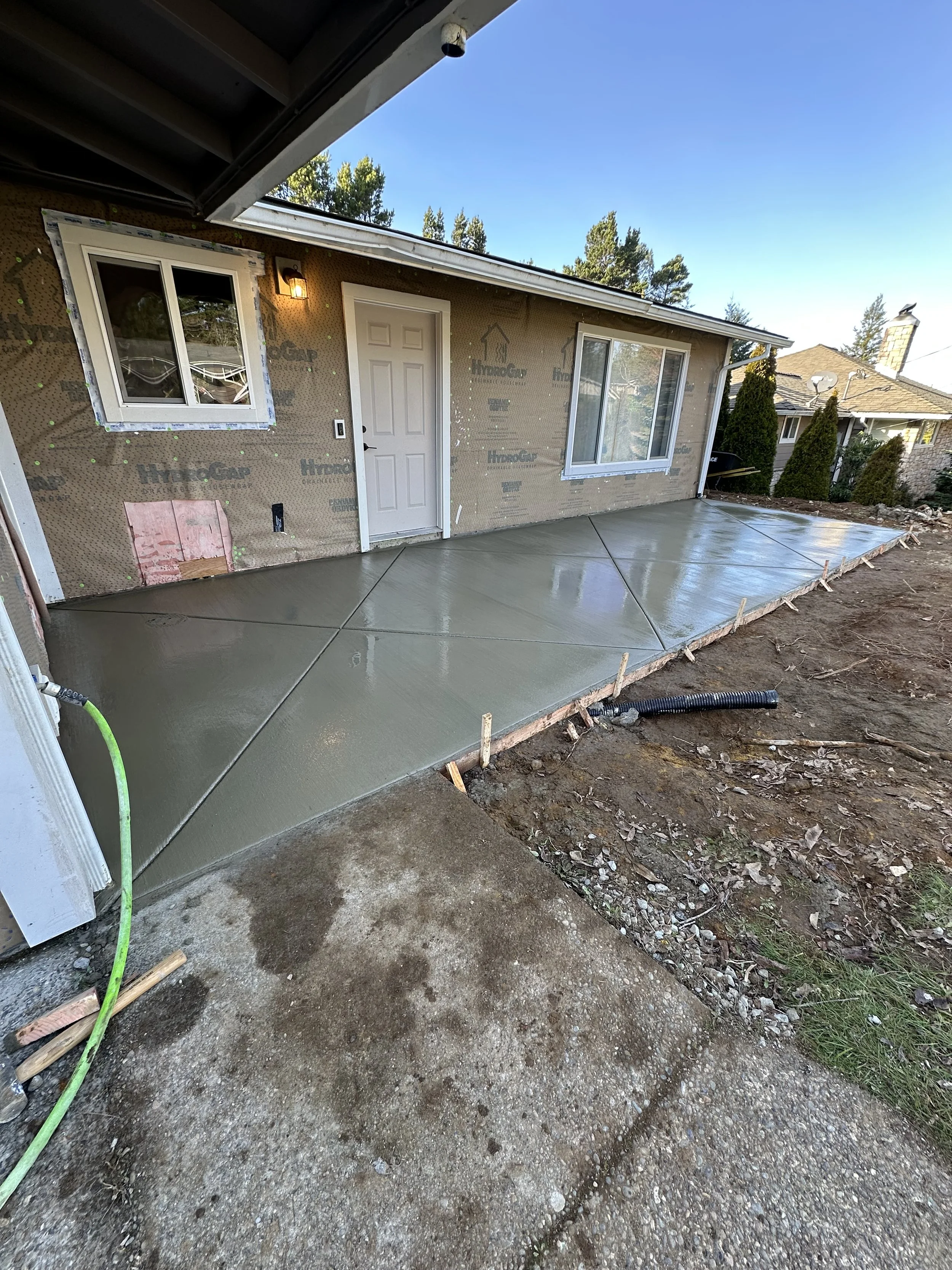 Freshly poured concrete patio in front of house with sliding glass door and window, construction materials and dirt surrounding the area.