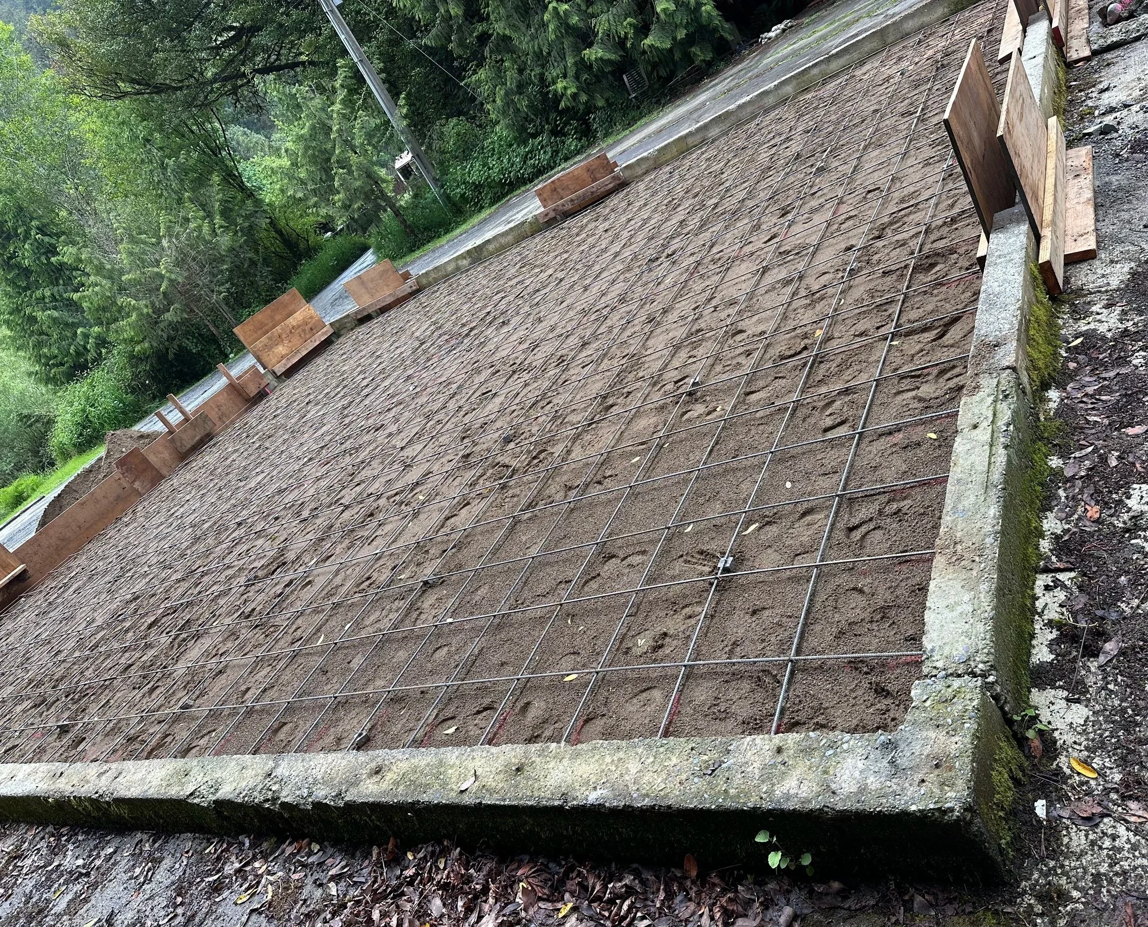 A construction site showing a foundation with steel rebar arranged in a grid pattern, surrounded by wooden formwork blocks, on a concrete base, in a wooded area with trees and greenery in the background.