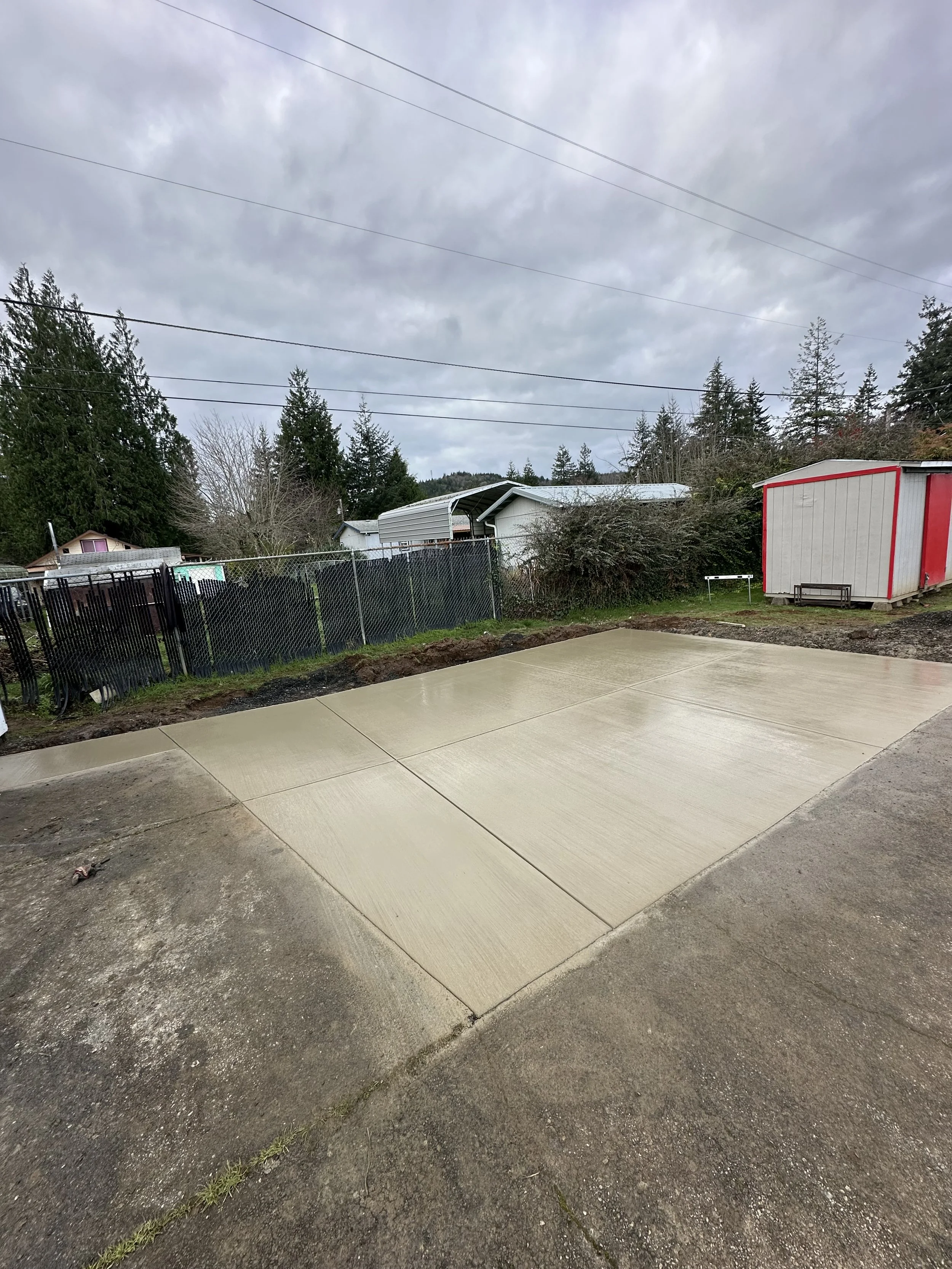 A concrete driveway with freshly poured sections, partially wet, next to an older asphalt surface. In the background, there is a chain-link fence, a shed with red trim, trees, and cloudy sky.