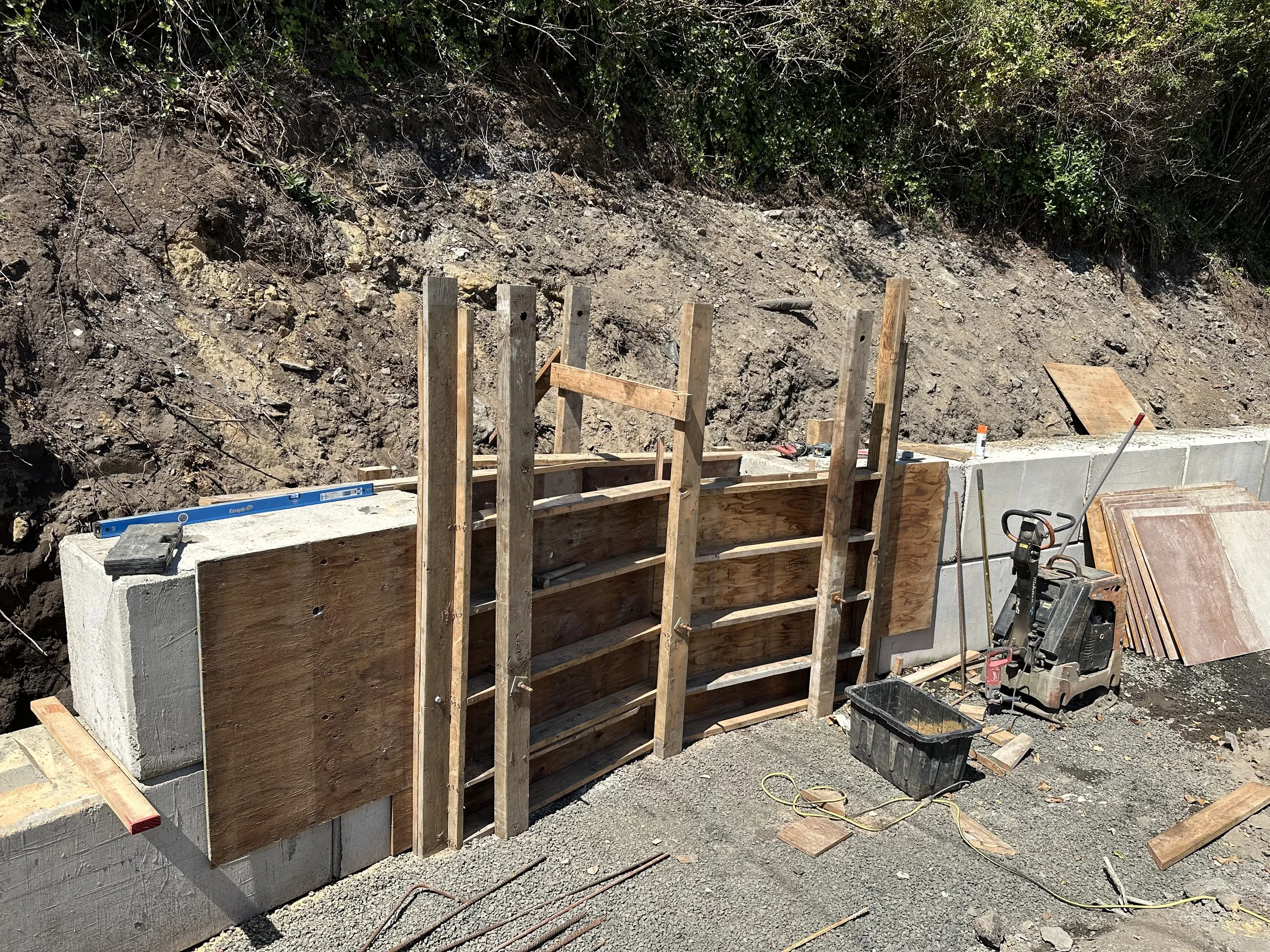 Construction site with wooden framing, concrete wall, tools, and a level on top, set against a dirt and grassy hillside.