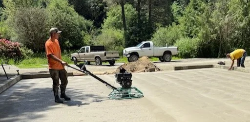 A person operating a compactor on a freshly poured concrete surface outdoors, with another worker in the background and trees in the background.
