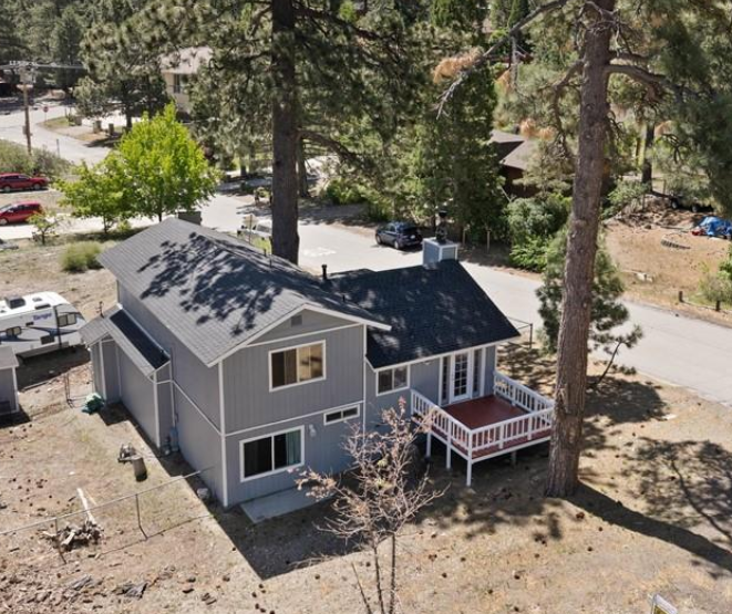 A gray two-story house with a small porch, surrounded by tall trees and some sparse vegetation, located in a suburban neighborhood.