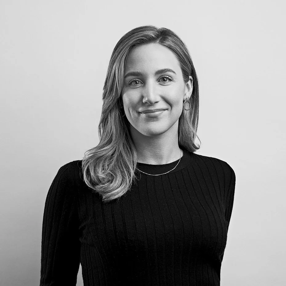 Black-and-white portrait of a young woman with shoulder-length hair, wearing a dark ribbed top, earrings, and a delicate necklace, smiling at the camera.
