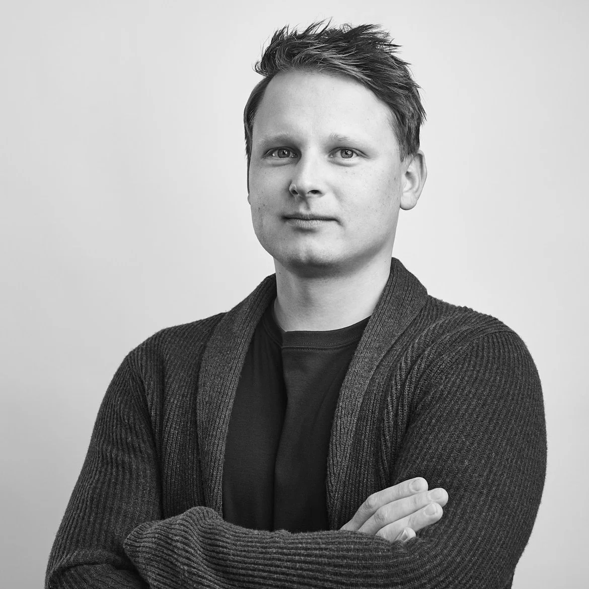 Black and white portrait of a young man with short, slightly messy hair, wearing a dark t-shirt and a knit cardigan, standing with arms crossed and looking at the camera against a plain light background.