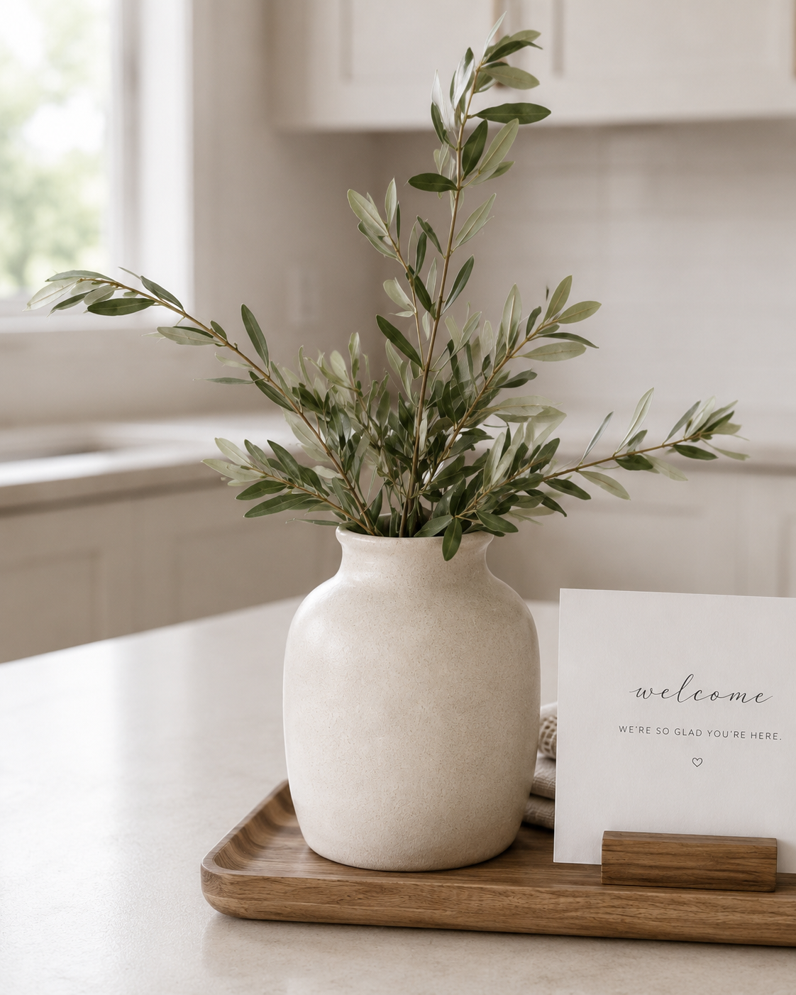 A beige ceramic vase with green leafy branches on a wooden tray, next to a welcome card with message 'we're so glad you're here' on a white surface.