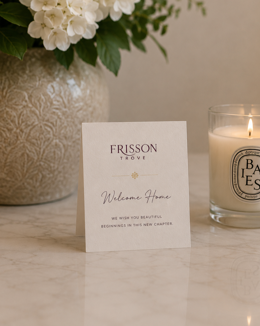 A white welcome card with elegant purple and black text on a marble surface, surrounded by a large flower arrangement in a decorative vase and a lit candle in a glass holder.