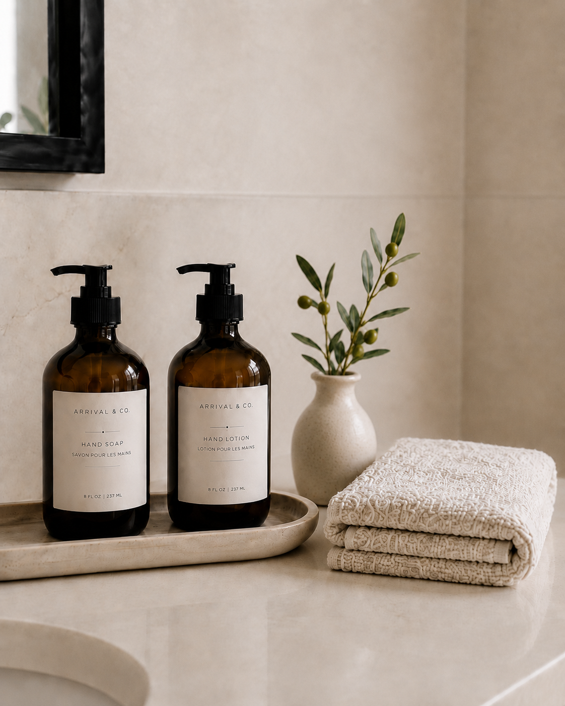 A bathroom countertop with two amber bottles labeled 'Arrival & Co' for hand soap and lotion, a small vase with greenery, and folded towels.