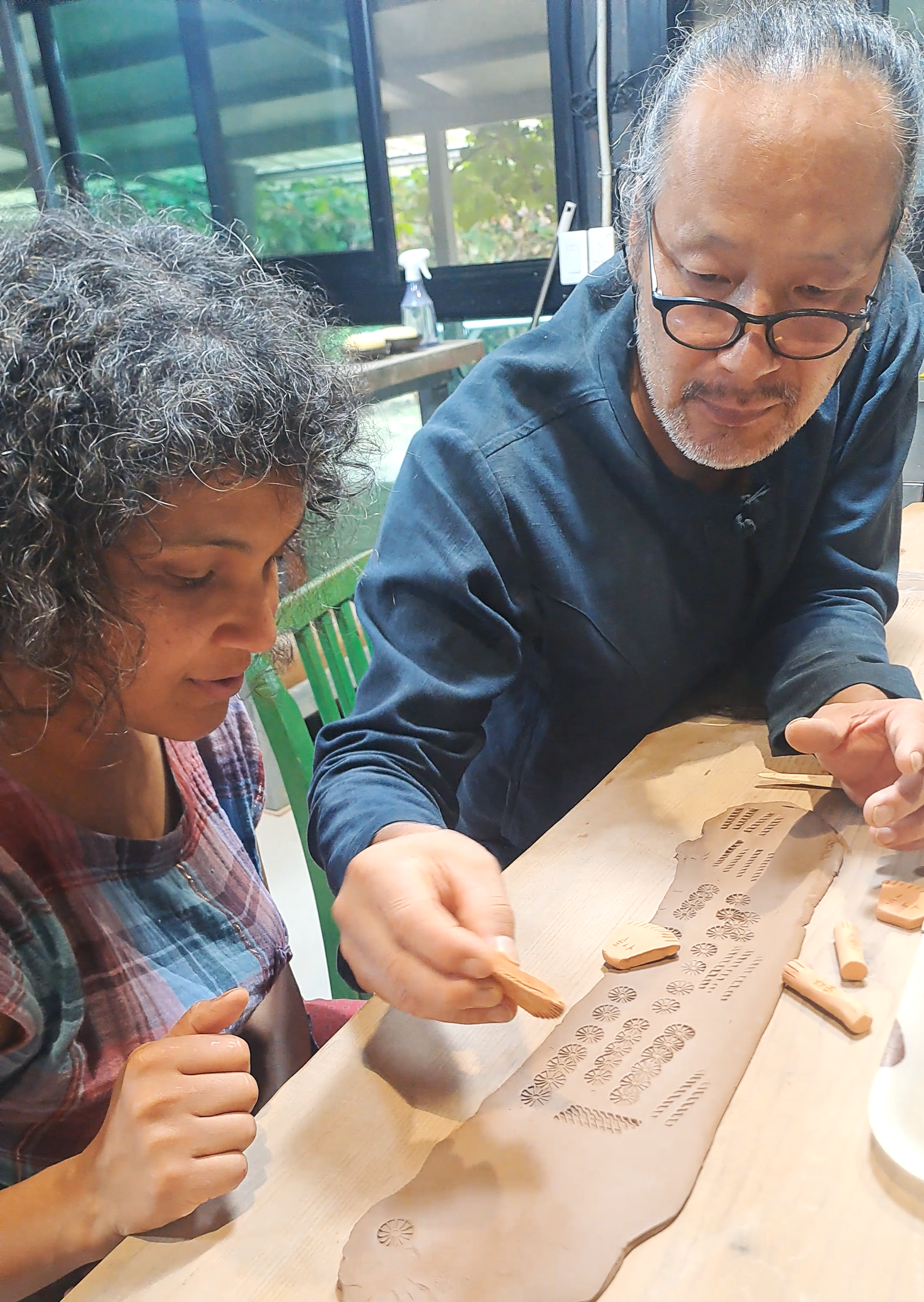 Two people working on clay sculpting at a wooden table with carved clay pieces and tools, in a room with large windows and green chairs.