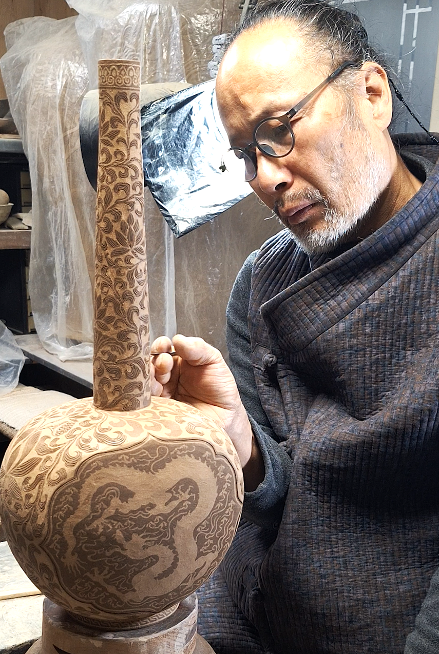 A man carving a wooden vase with intricate dragon and floral patterns in a woodworking studio.