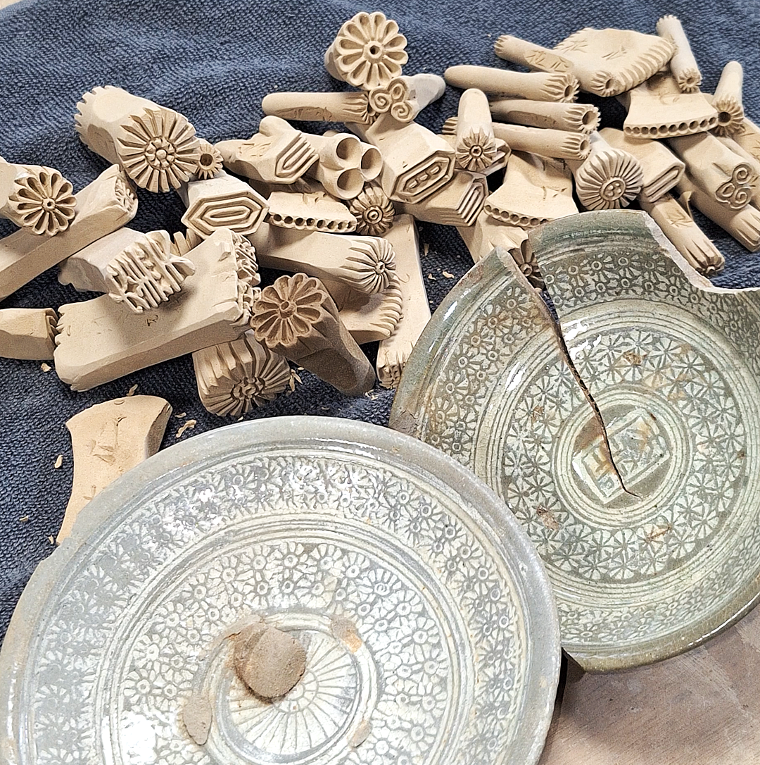 Two broken glass plates with intricate patterns and a pile of carved wooden stamps with various designs on a dark fabric surface.