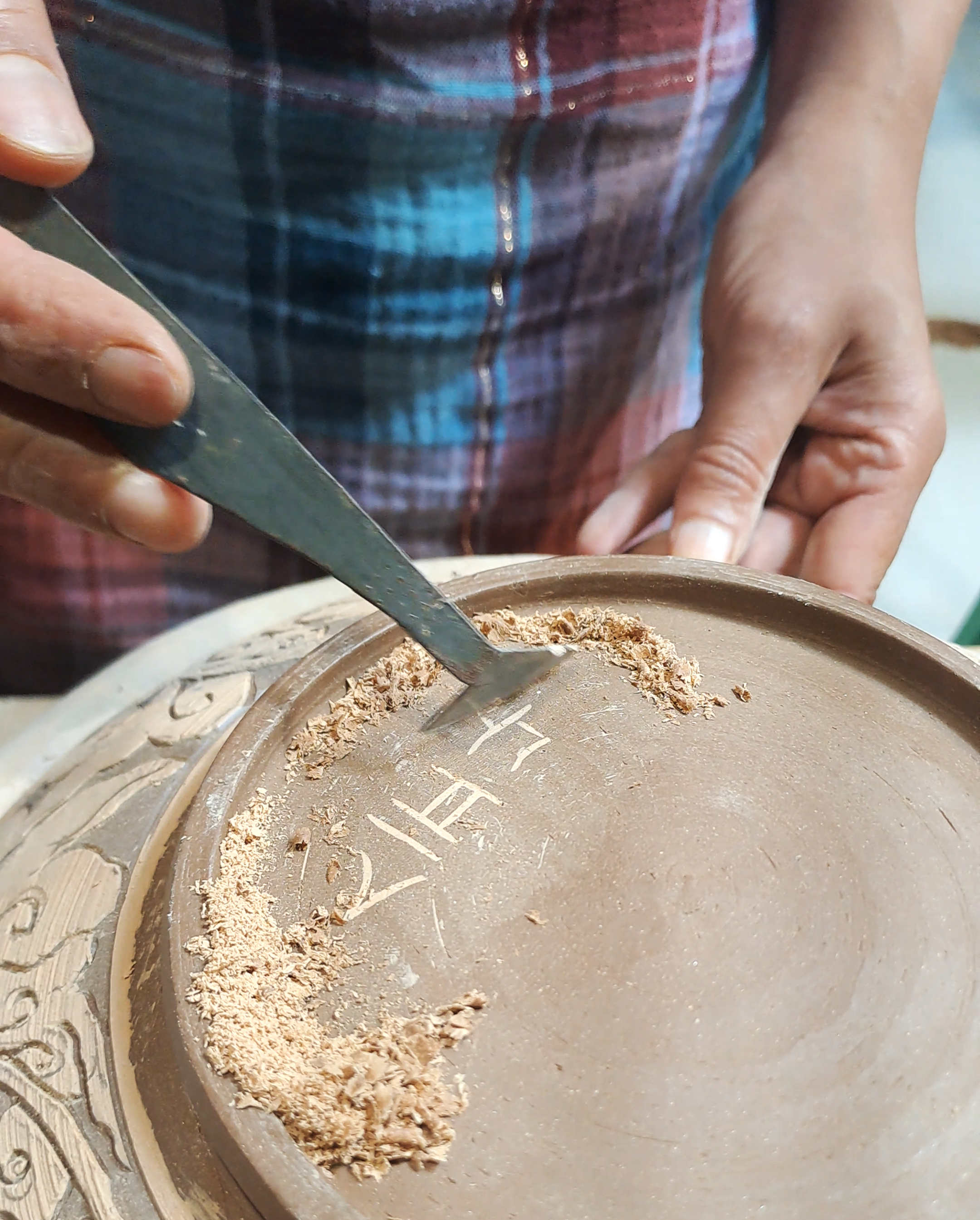 A person wearing a plaid shirt is using a carving tool to scrape or carve something on a round, brownish surface, possibly clay or ceramic. The focus is on their hands and the surface they are working on.