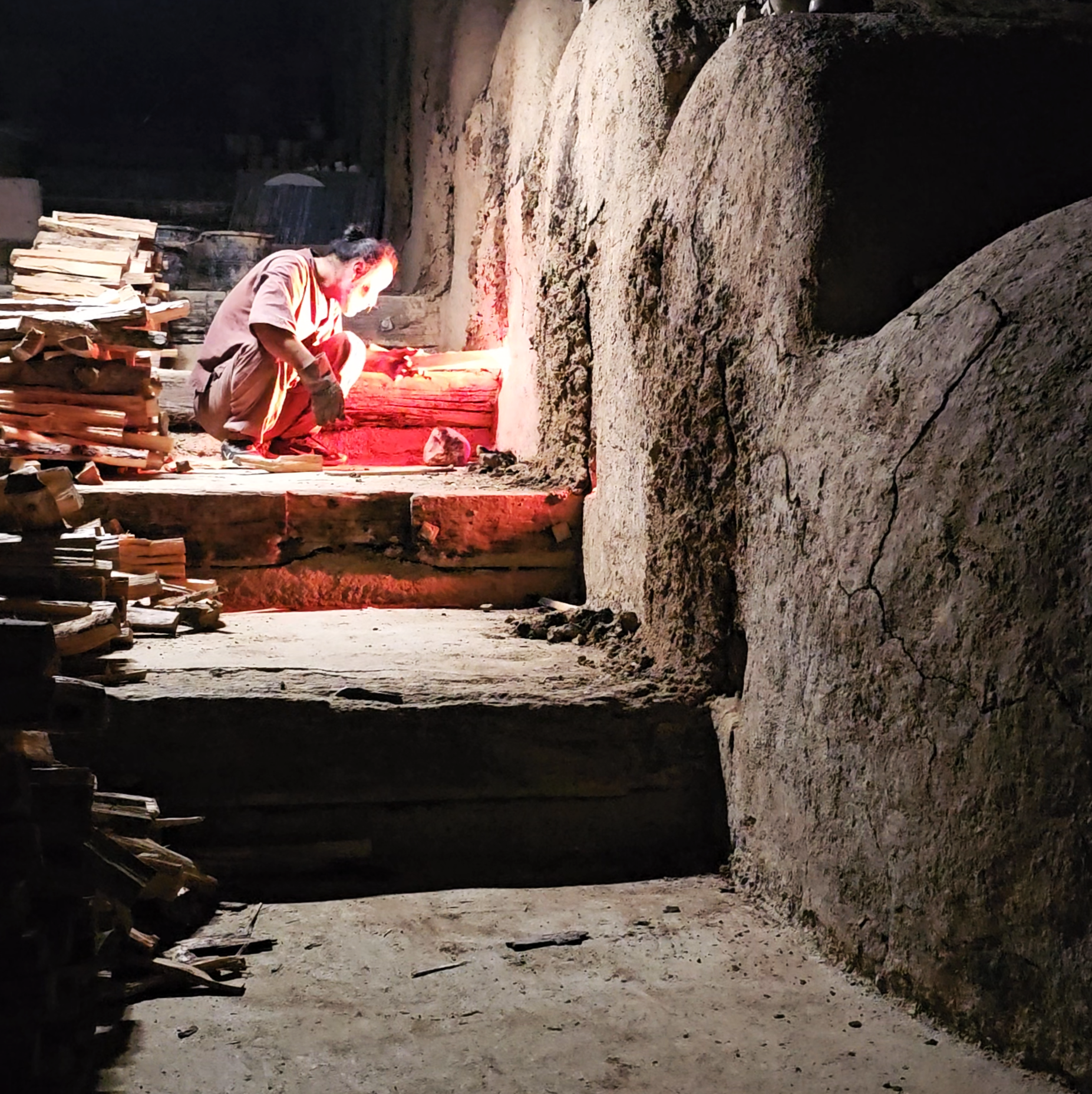 Person kneeling in a dimly lit stone-walled cellar or cave, tending to a small fire with logs nearby.