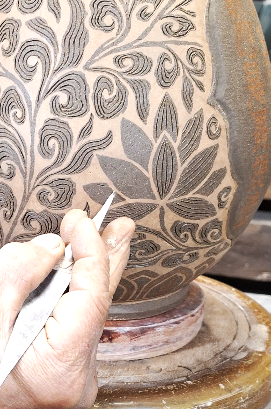 A person carves intricate floral and swirl patterns into the surface of a large clay or ceramic vessel using a small tool.