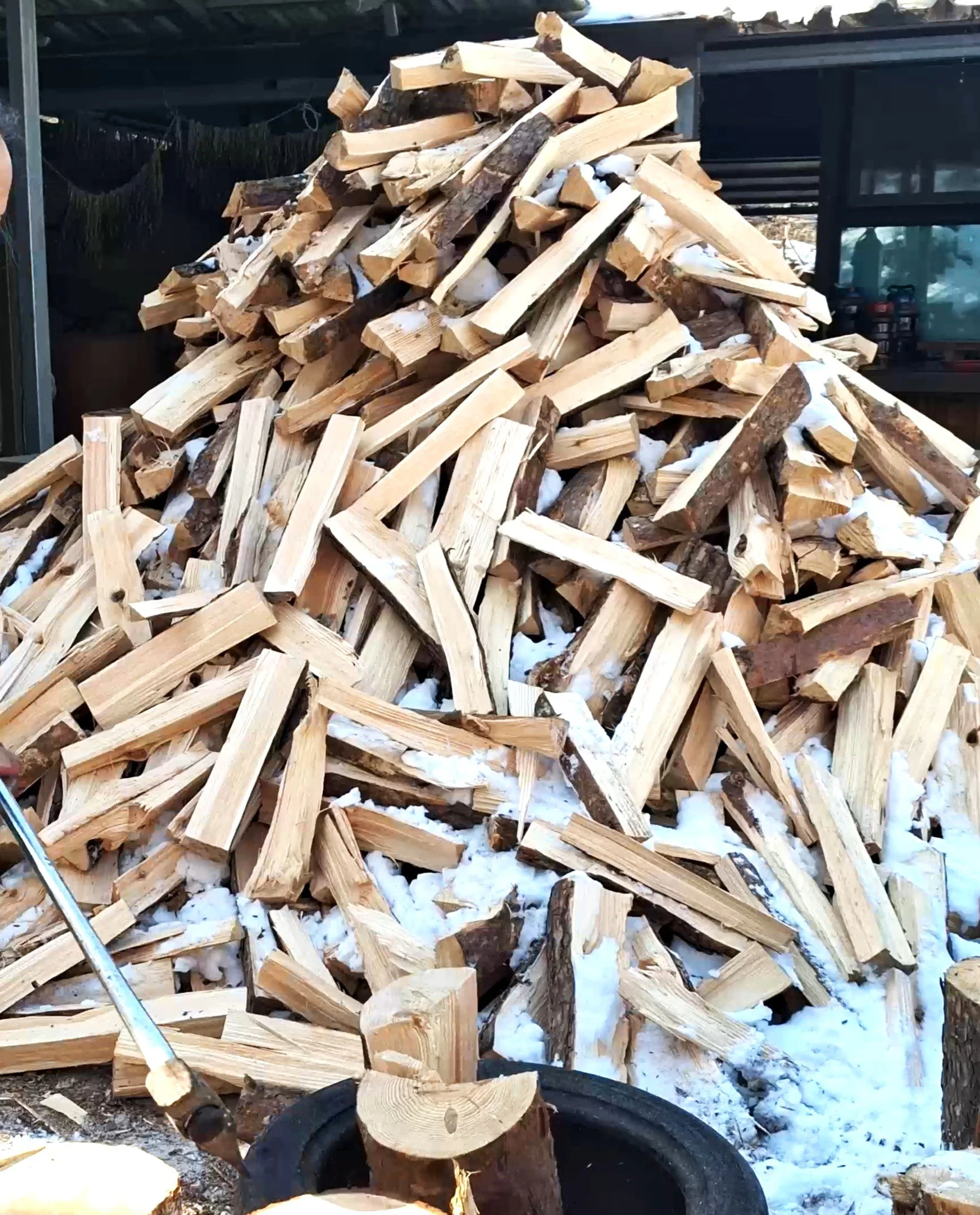 A large pile of split firewood stacked outdoors with snow on the ground, with some chopped logs visible at the bottom and a black container around the base.