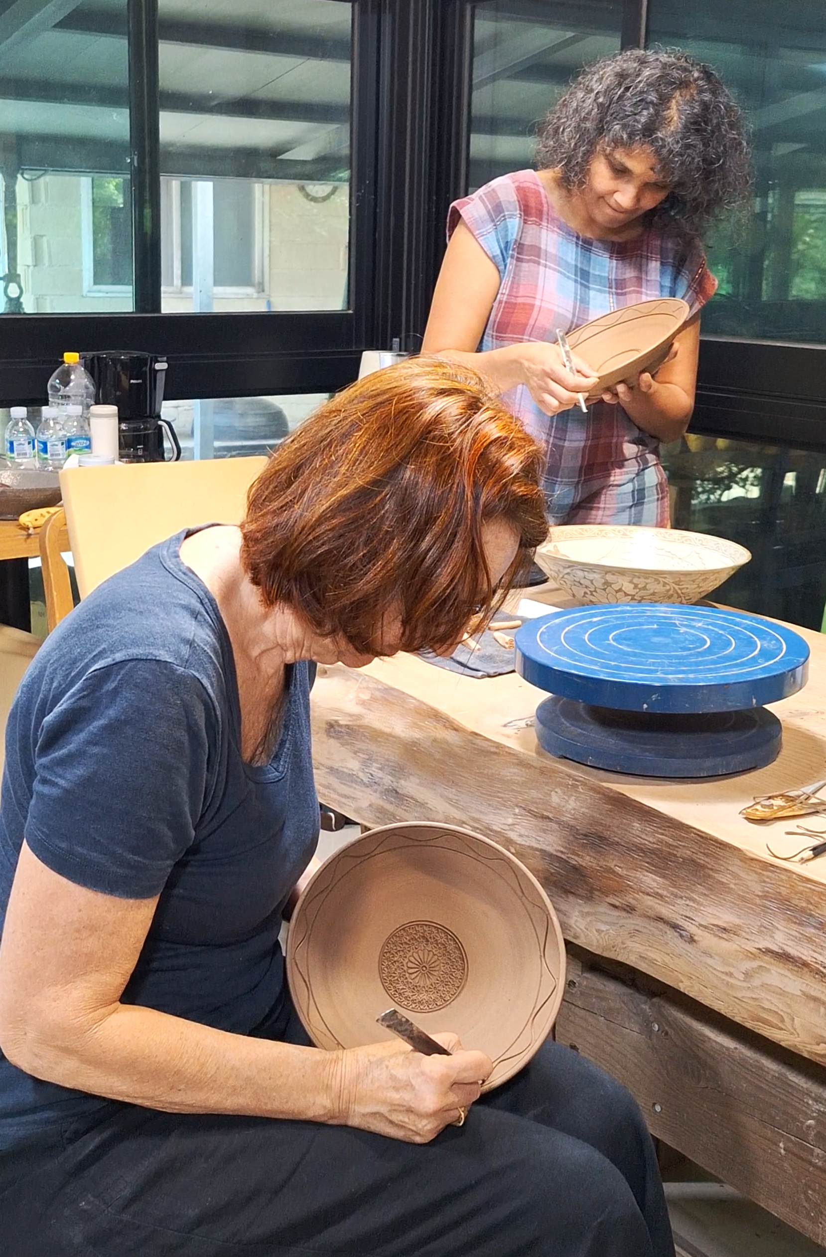 Two women are working on decorating or painting large pottery plates in a workshop. One woman with gray curly hair is standing and writing or drawing on a plate, while the other woman with red hair is seated, holding a decorated plate and working on its design.