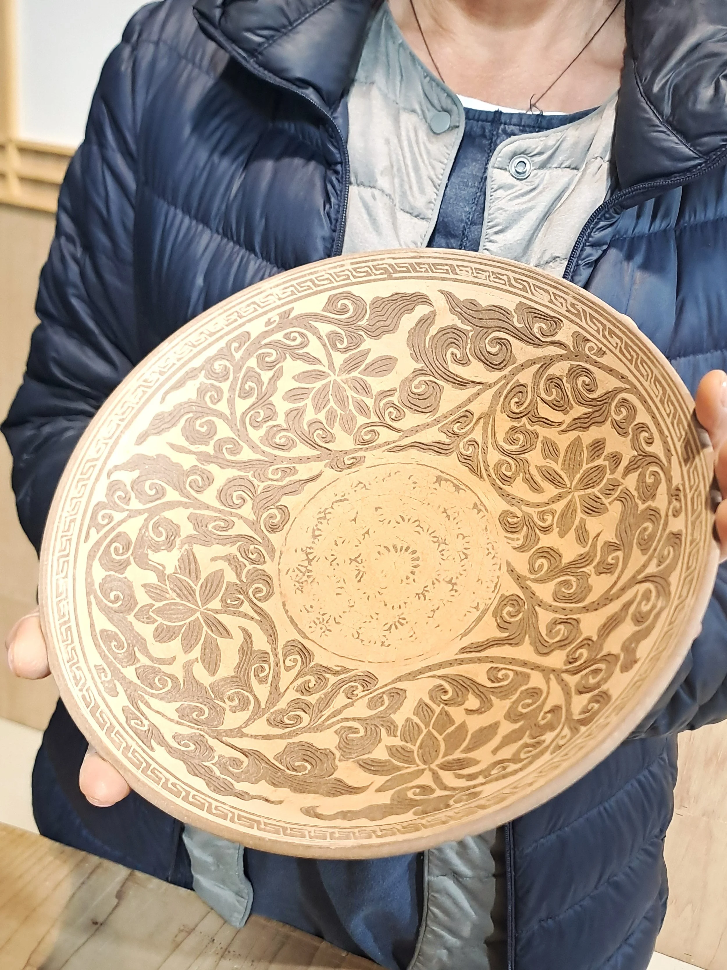 Person wearing a dark puffy jacket and gray shirt holds a round decorative wooden tray with intricate carved floral and swirl patterns.