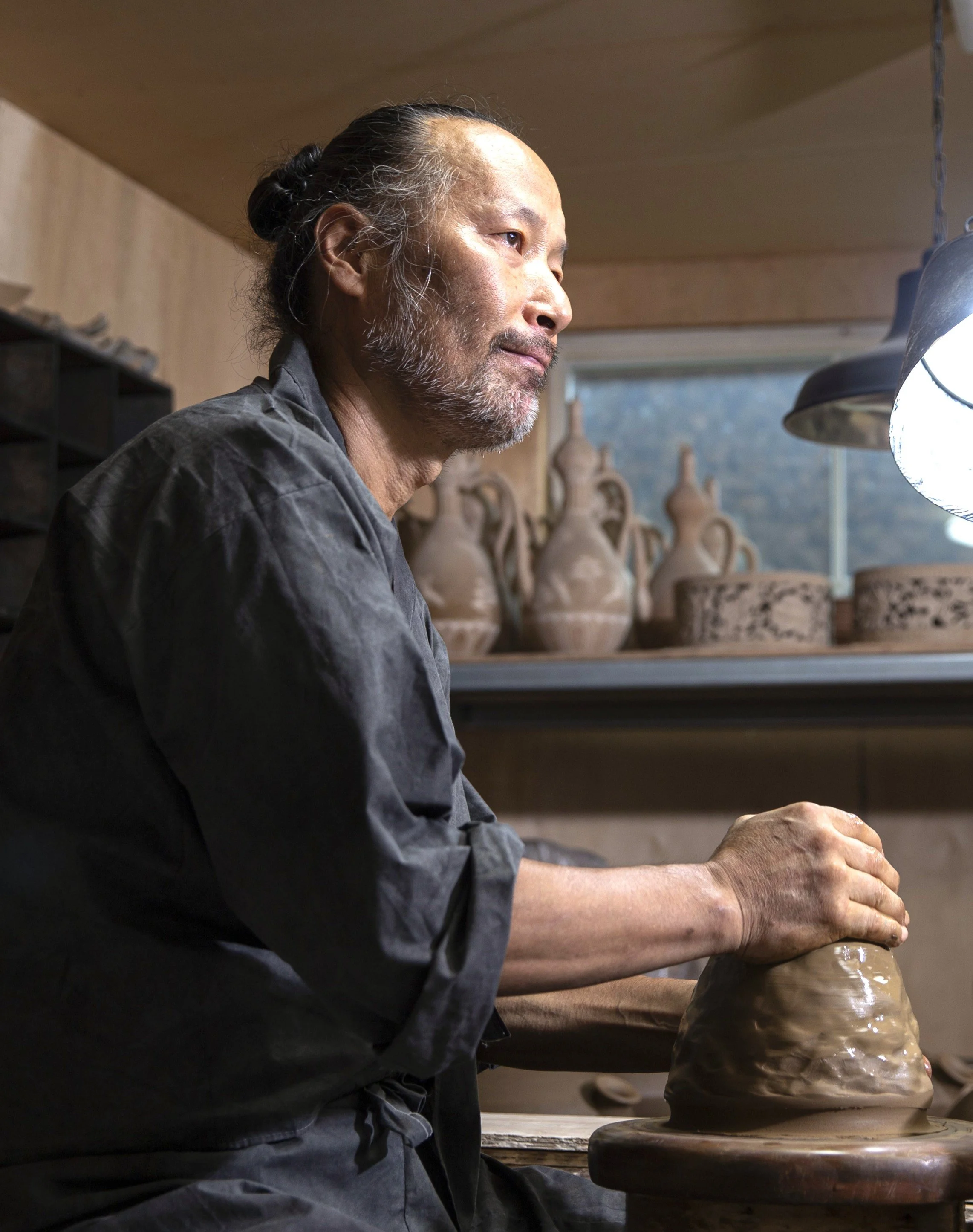 An elderly man with gray hair and beard, wearing a black shirt, working on a pottery wheel shaping a clay pot in a ceramics studio, with shelves of finished pots in the background.