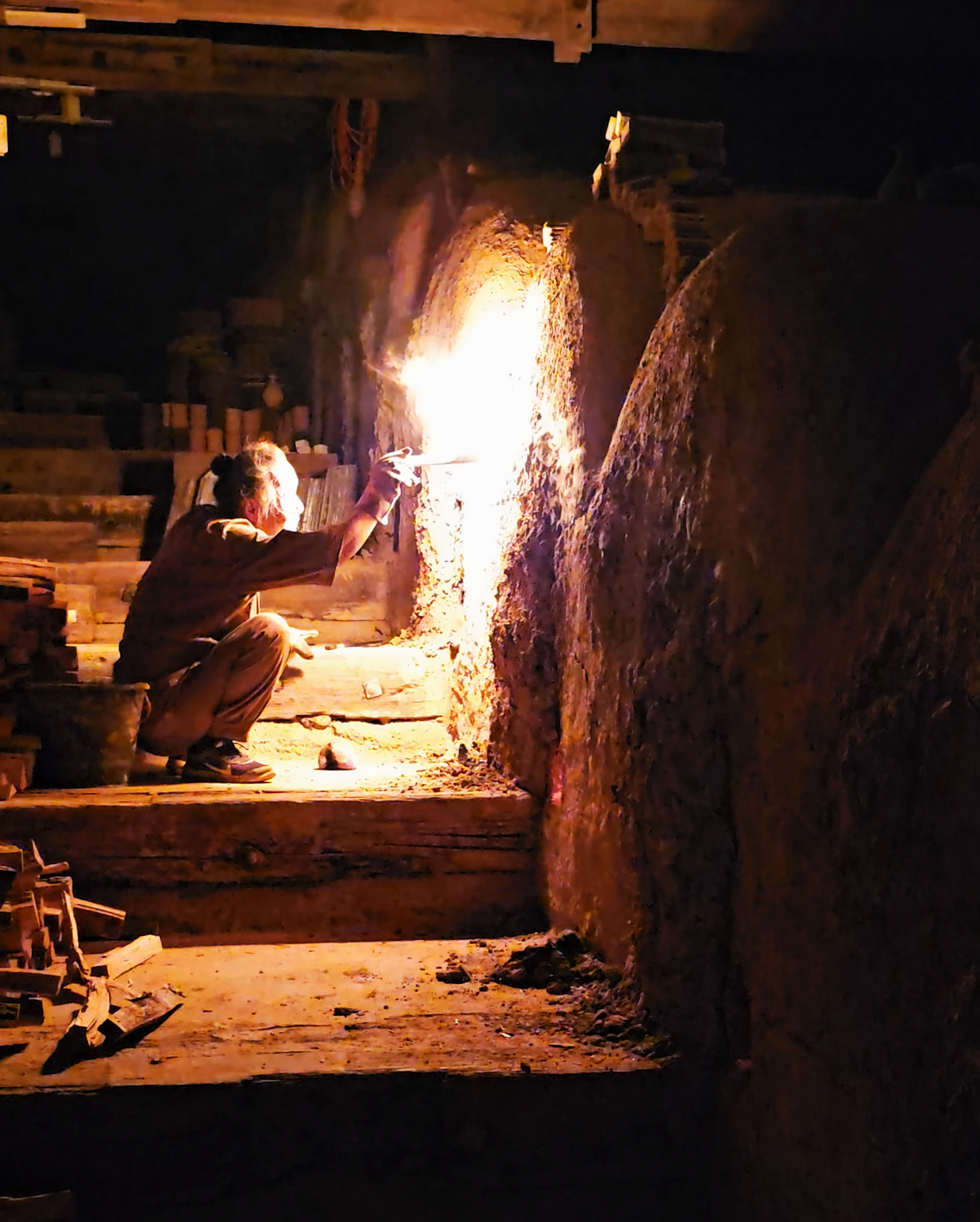 A person wearing gloves kneeling inside a dark tunnel or cave and working on a large vertical molten metal stream or lava flow, with bright orange and yellow flames illuminating the scene.