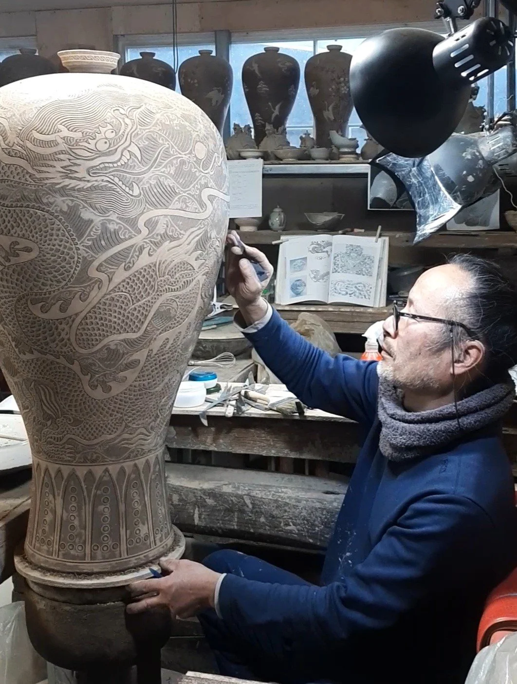 An artist working on a large ceramic vase with intricate dragon carvings in a pottery studio.