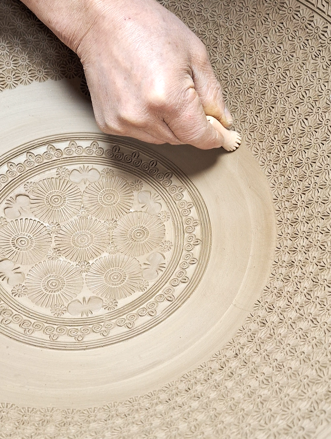 Close-up of a hand carving intricate patterns into a ceramic or clay surface, with detailed floral and geometric designs.