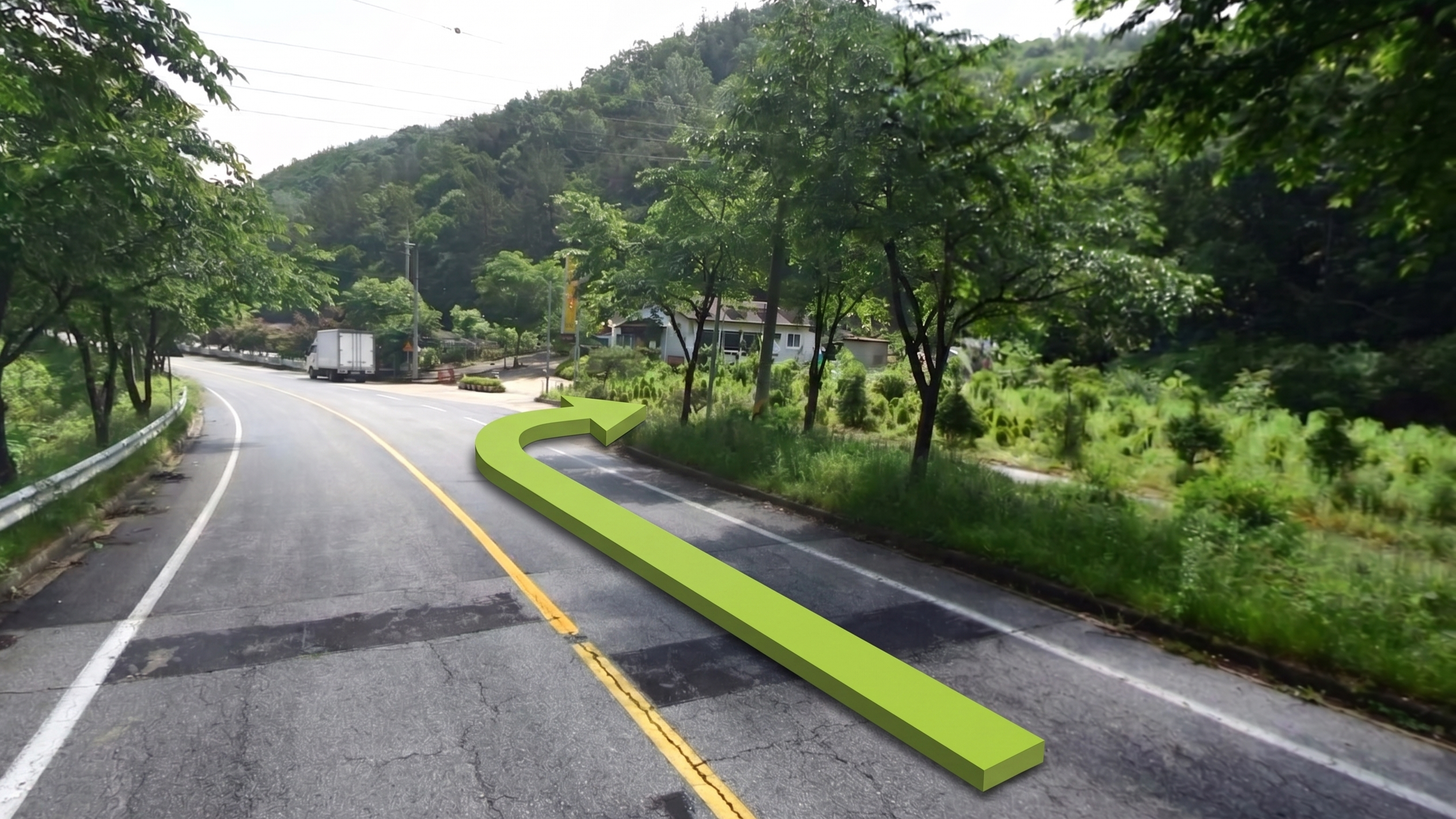 A winding road surrounded by lush green trees and hills. There is a green arrow on the road indicating a right turn. A few houses are visible in the background along with utility poles and wires.