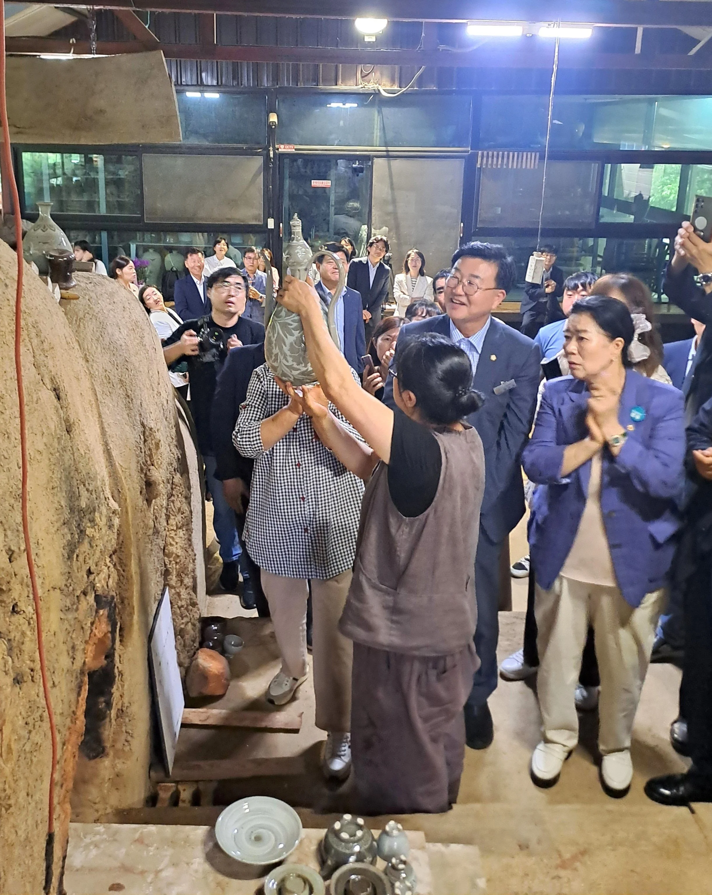 Group of people in a pottery or ceramics workshop, looking at a large clay or ceramic piece. Some individuals are taking photos or videos, and a display of ceramic objects is visible in the foreground.