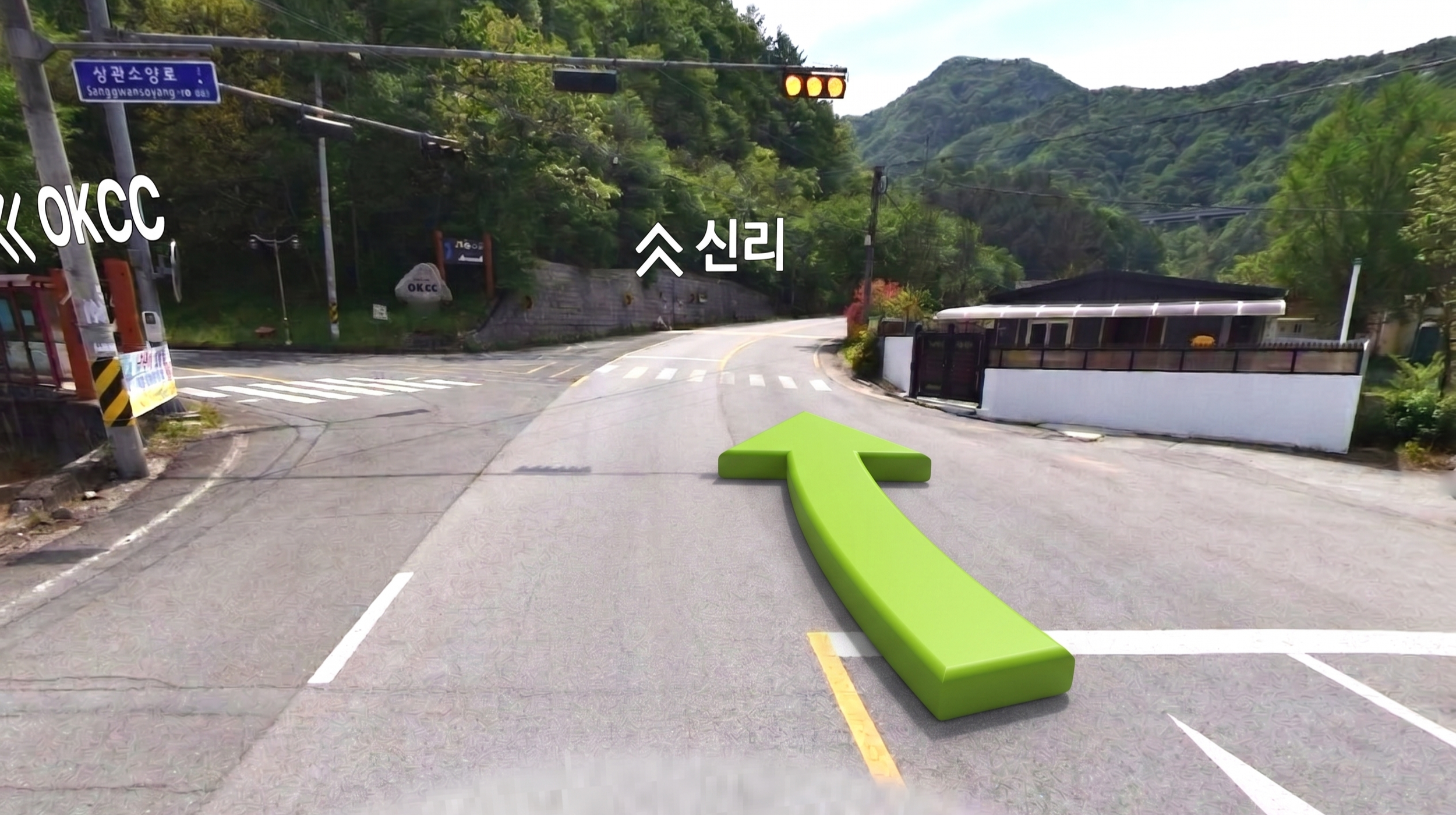 A mountain road with directional signs and a large green arrow pointing straight ahead. One sign is blue with white Korean text and English translation indicating the direction to Sanggwansoyang. The scene includes lush green mountains and a small structure on the right side. There are traffic lights and electrical wires overhead.