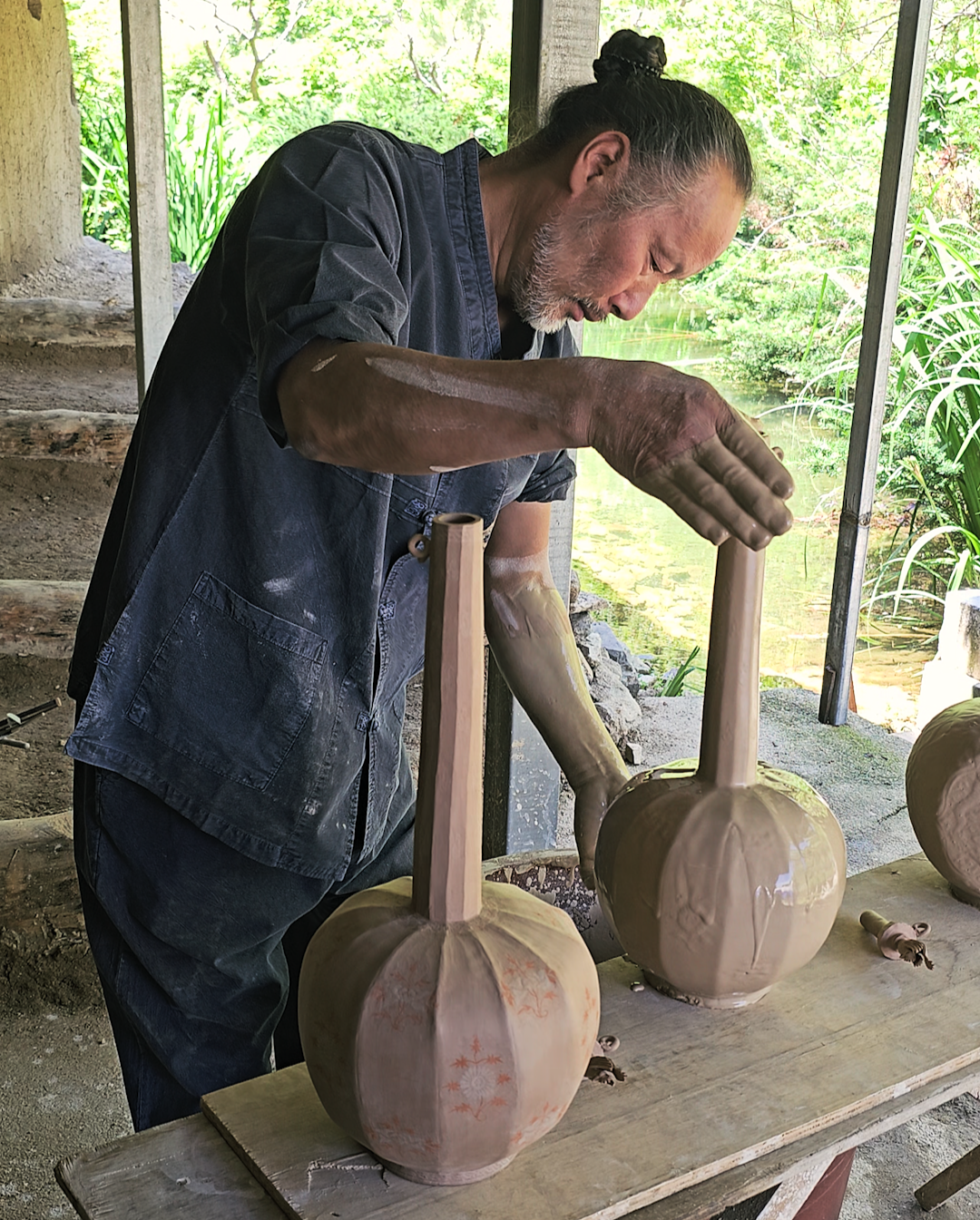 A man with long hair tied in a bun, wearing a dark blue work apron, is working on a handcrafted pottery piece outdoors. The pottery appears to be in the early stages of creation, shaped and drying on a workbench with a natural backdrop of green folia