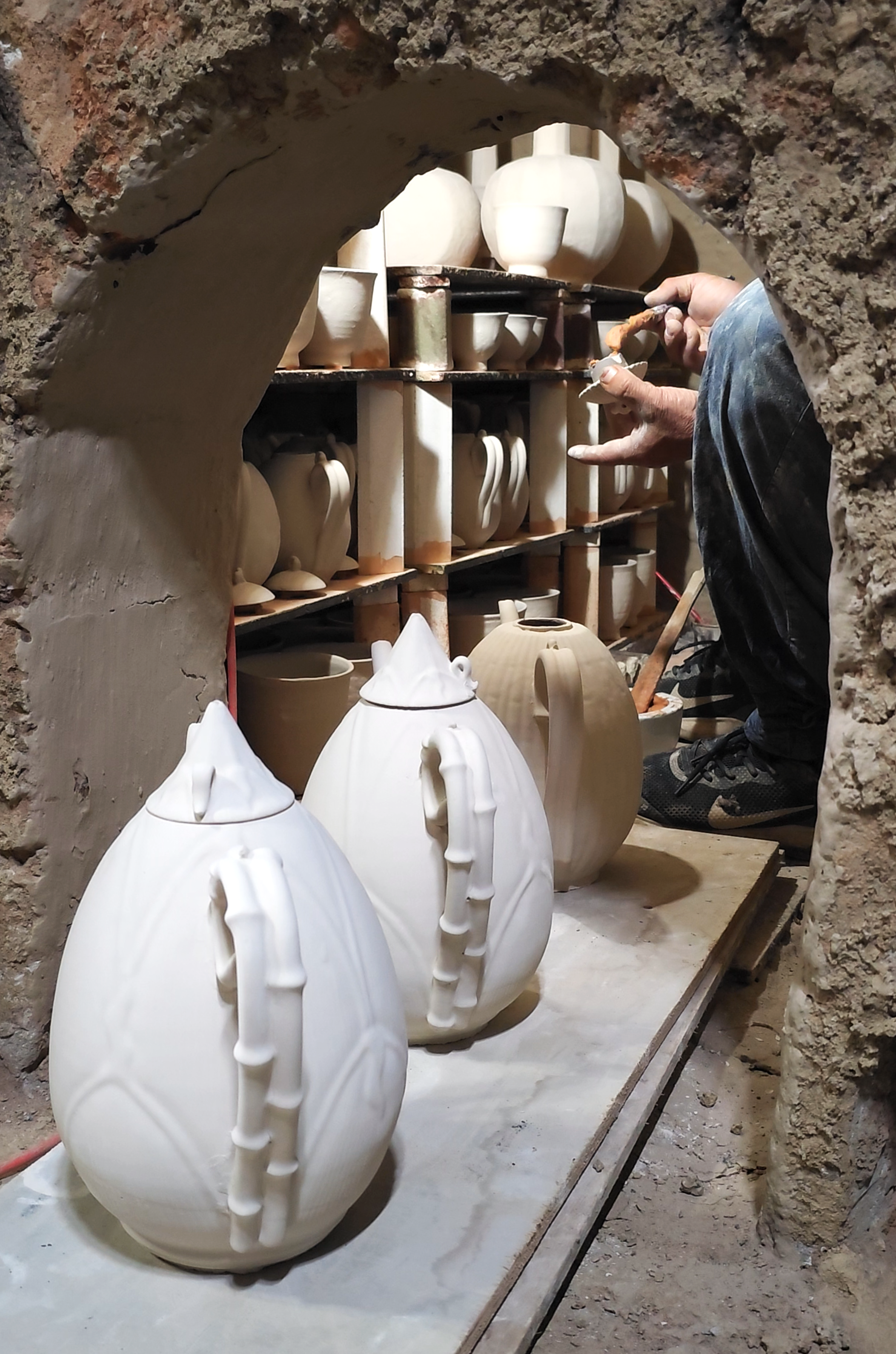 A person working on creating ceramic vases and teapots inside a pottery workshop, viewed through a rough brick archway.