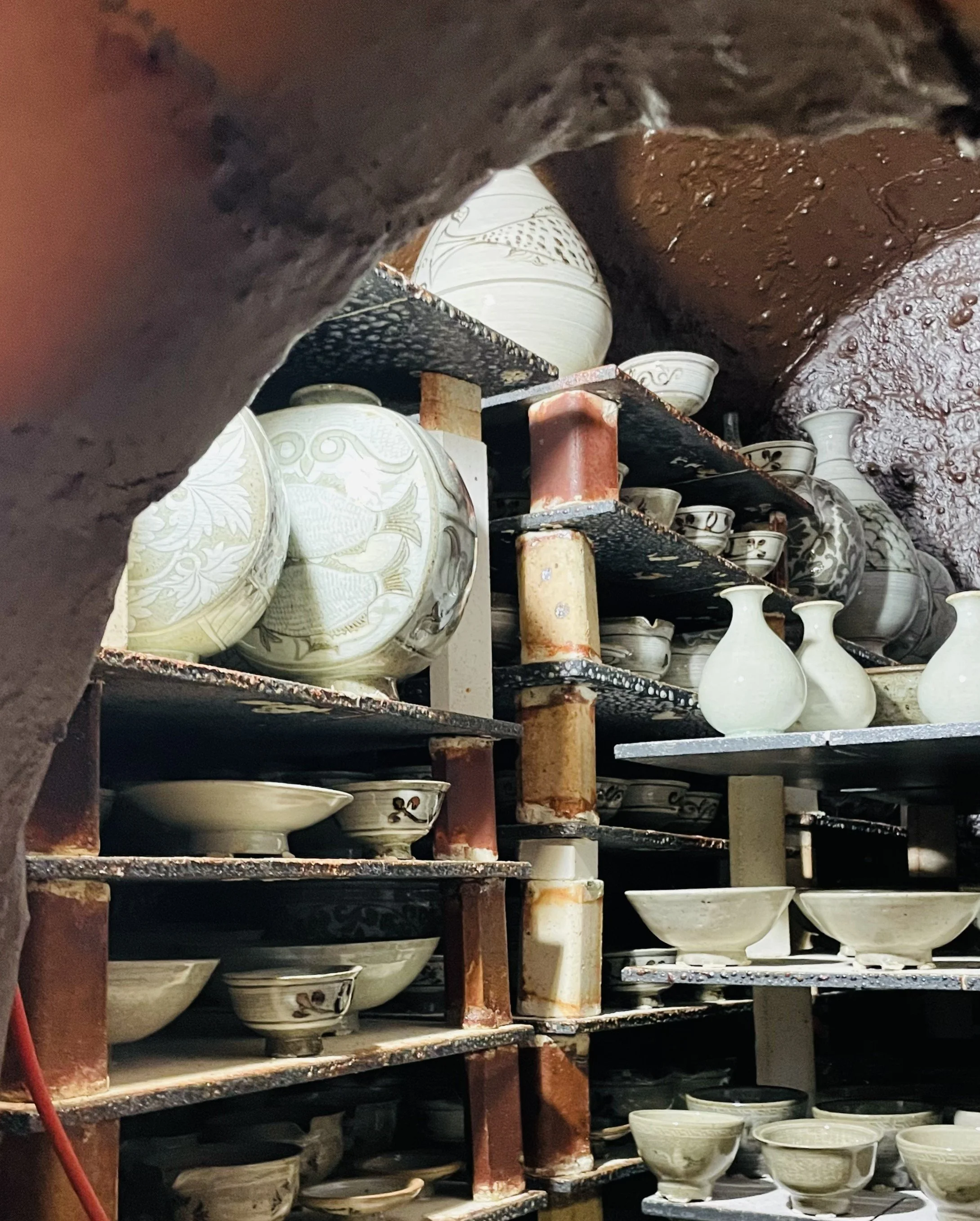 A storage shelf filled with multiple white ceramic bowls featuring intricate gold or gray patterns, situated in a dark and rusty space with metal and pipe structures.