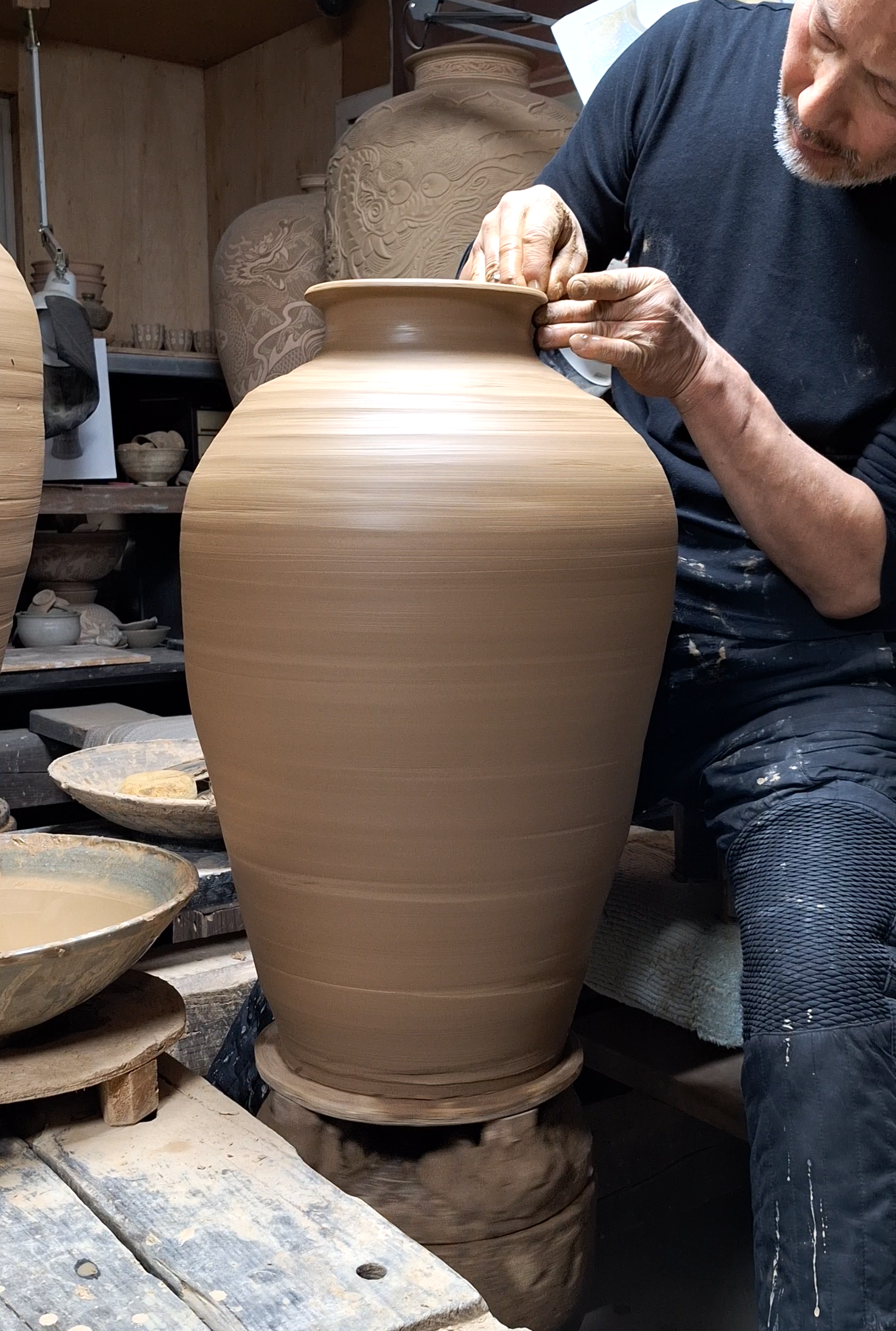 A man shaping a large clay vase in a pottery studio, with shelves of finished pottery and tools in the background.