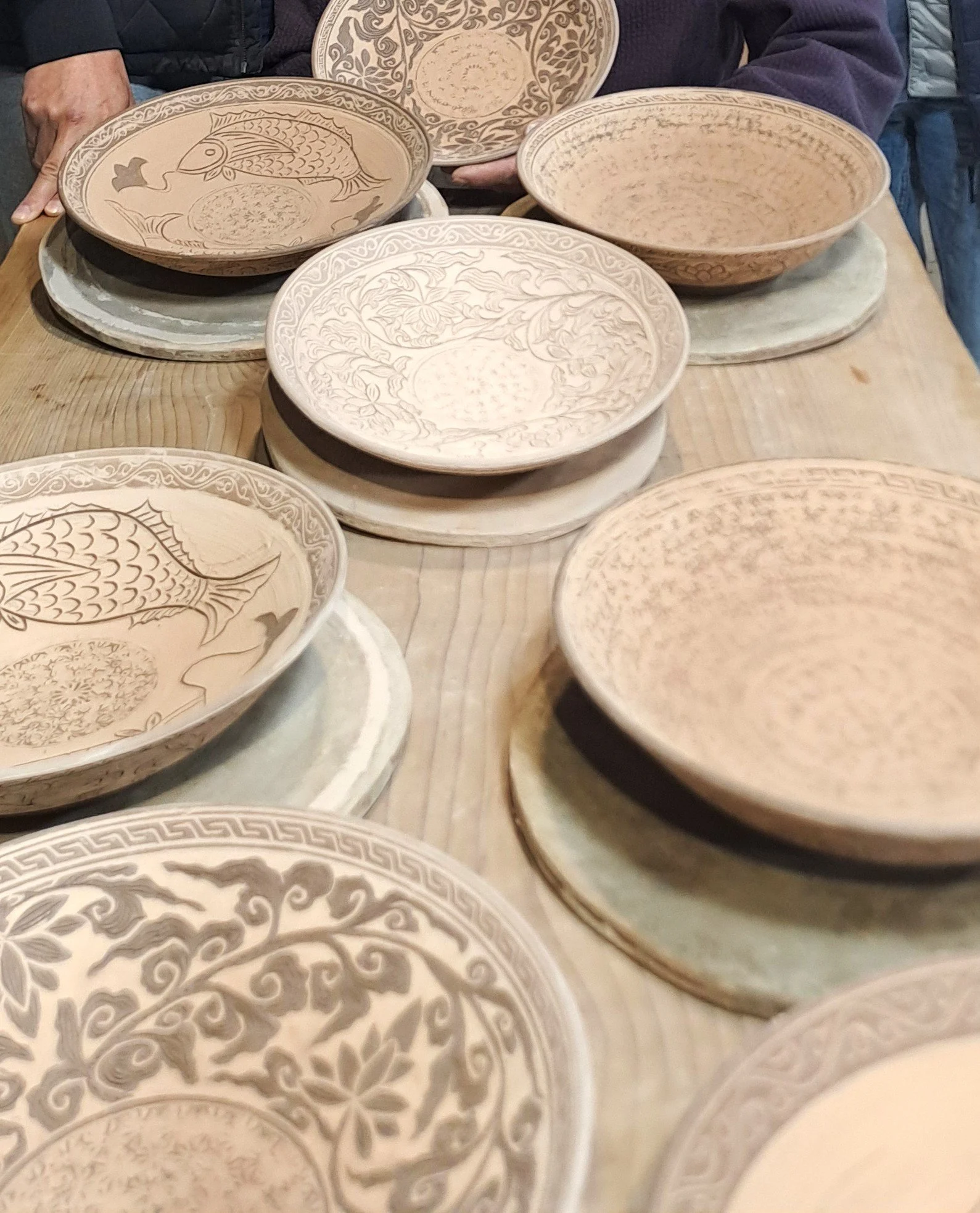Several decorated oval and round ceramic bowls on a wooden table, some resting on white bases, with people in the background.