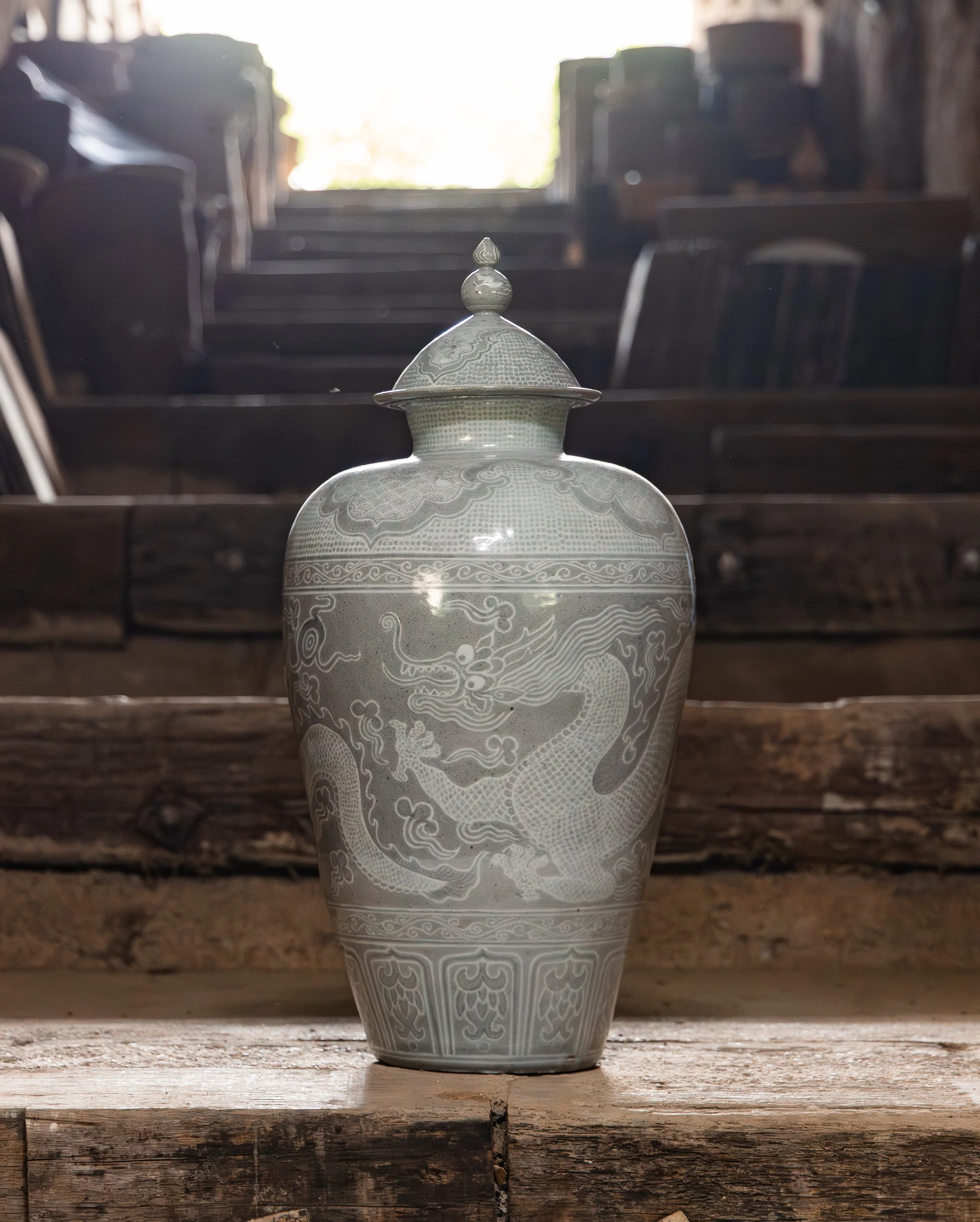 A white ceramic vase with intricate dragon designs, placed on a wooden surface. In the background, there are stacked chairs and a bright light source.